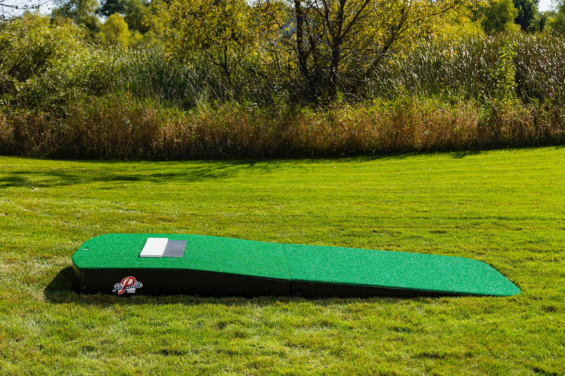 The Portolite Standard Two-Piece Practice Mound, a green, rectangular portable mound for all ages, sits on a grassy lawn with trees and tall grass in the background. Featuring an angled surface and designed to resemble a traditional pitching mound, it’s perfect for outdoor training or fun.