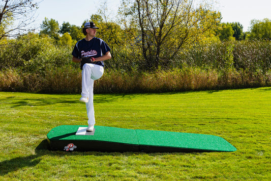 A baseball player in a navy jersey and white pants stands mid-pitch on the Portolite Standard Two-Piece Practice Mound, a portable mound for all ages, set on a grassy field with trees and bushes in the background under a clear sky.