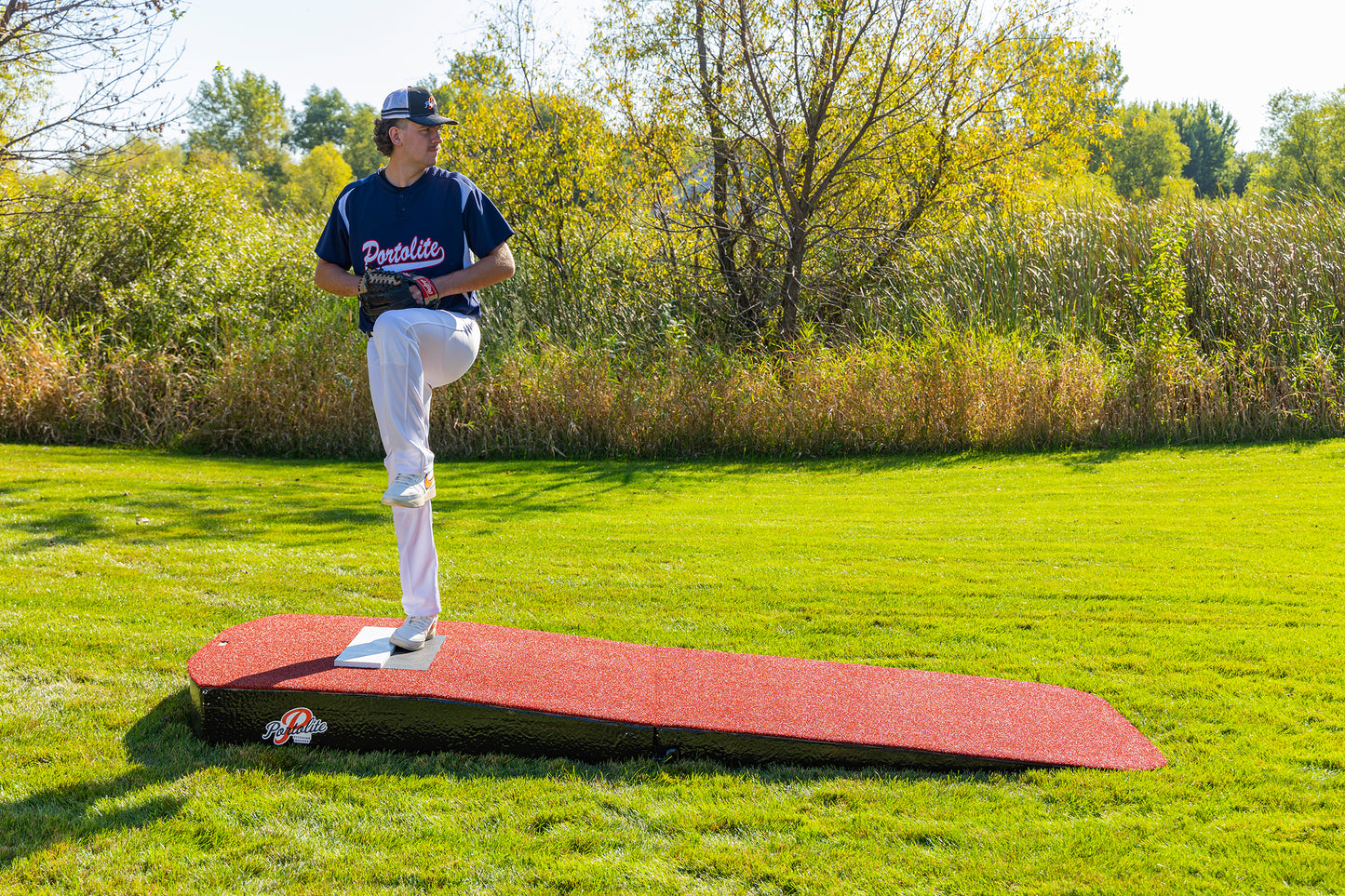 A baseball player in a blue shirt and white pants stands on the Portolite Standard Two-Piece Practice Mound, a portable mound designed for all ages, preparing to pitch in a grassy outdoor area with trees in the background.