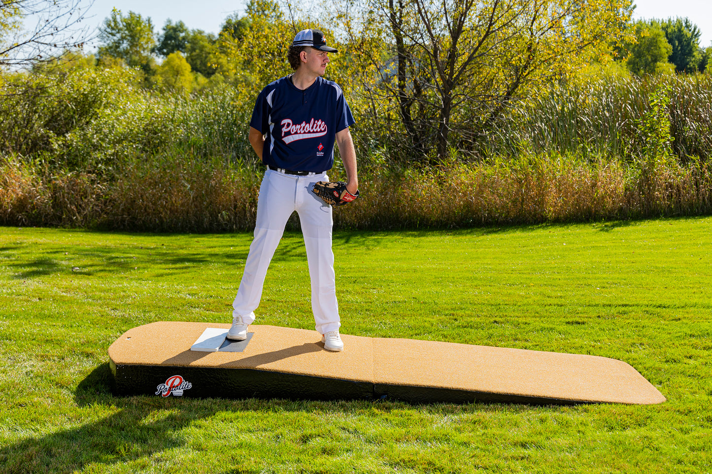 A baseball player in a navy jersey and white pants stands outdoors on the Portolite Standard Two-Piece Practice Mound | Portable Mound for All Ages, looking to the side, with green grass and trees in the background.