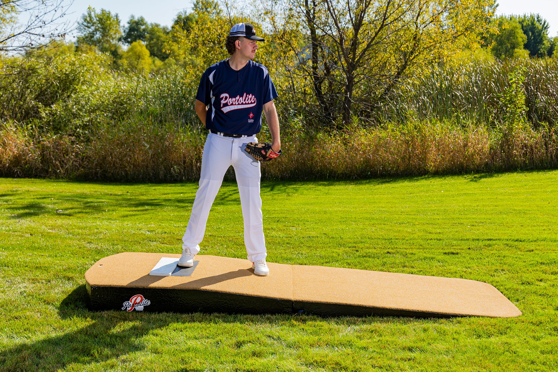 A baseball player in a navy jersey and white pants stands outdoors on the Portolite Standard Two-Piece Practice Mound | Portable Mound for All Ages, looking to the side, with green grass and trees in the background.