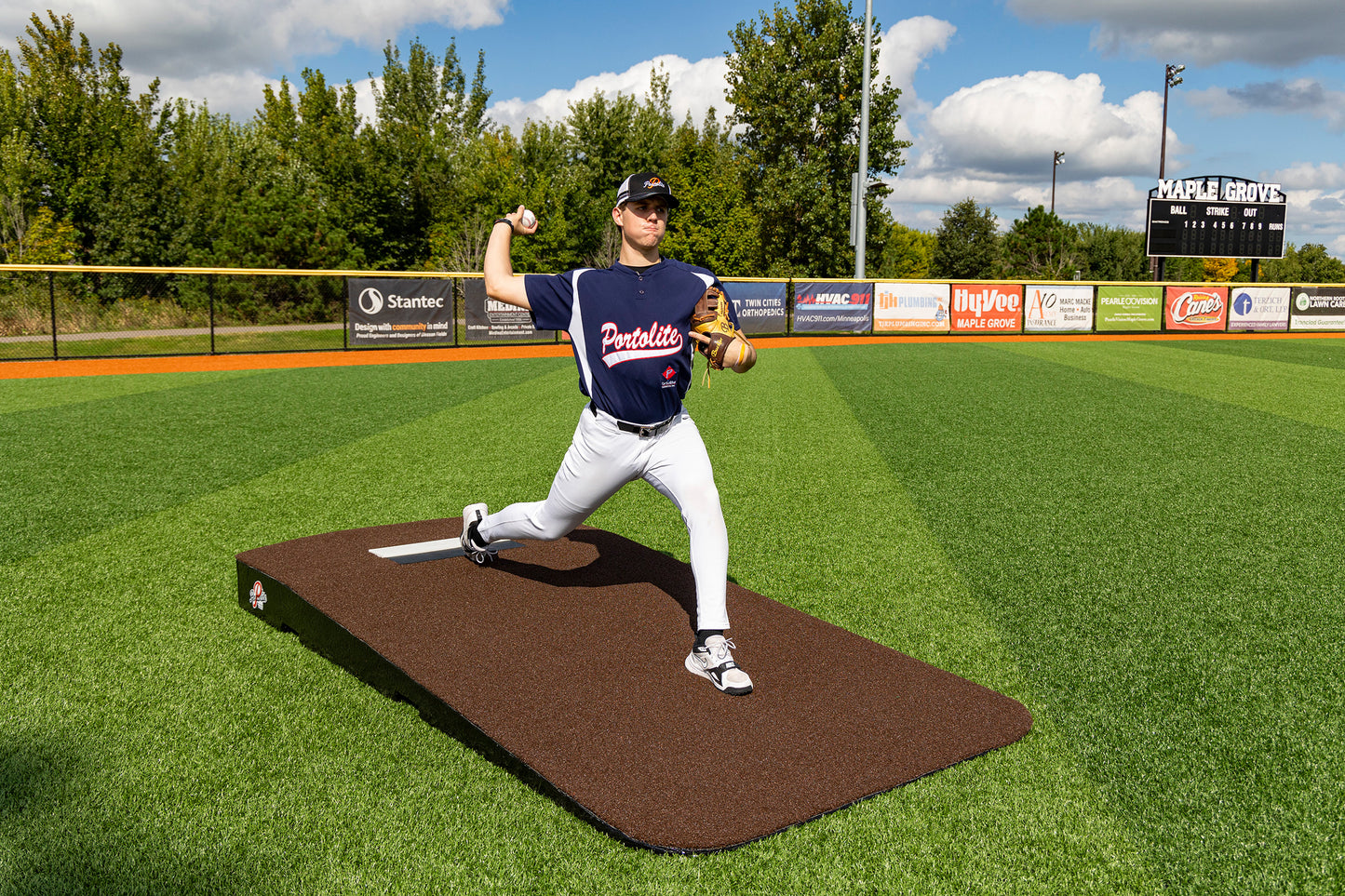 A baseball player in a blue jersey pitches from the Portolite Oversized One-Piece Practice Mound—designed for the longest stride for tall pitchers—on a green field surrounded by trees, an electronic scoreboard, and advertisements, creating an ideal training environment.