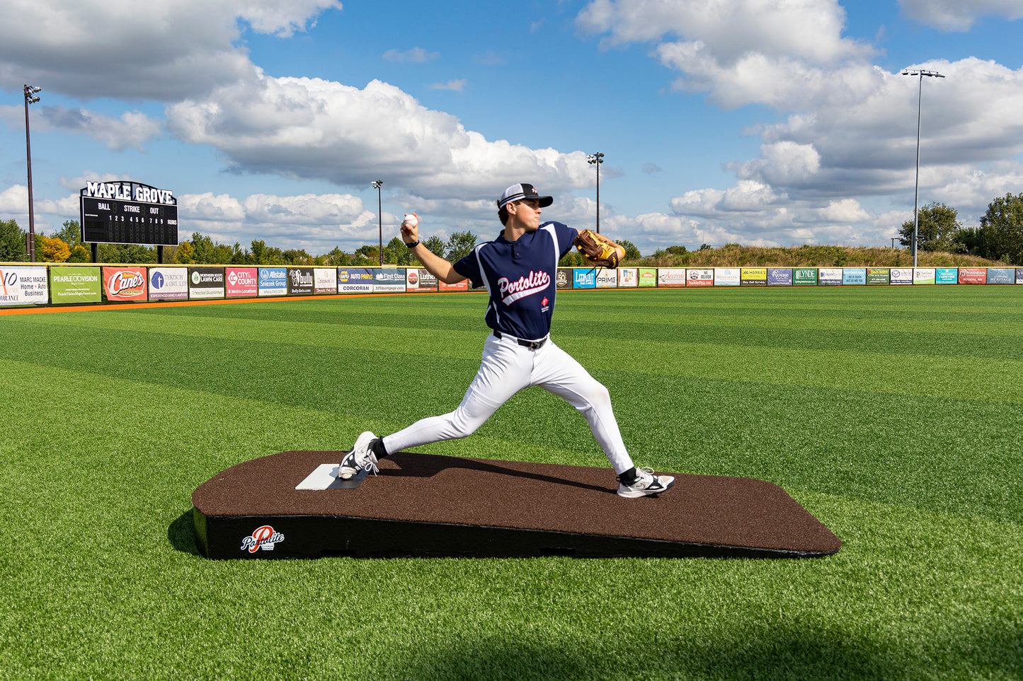 A baseball player in a blue jersey winds up to pitch on the Portolite Oversized One-Piece Practice Mound—designed for the longest stride for tall pitchers—on an empty field with a scoreboard and advertisements in the background under a partly cloudy sky, making it perfect for pitchers’ training.