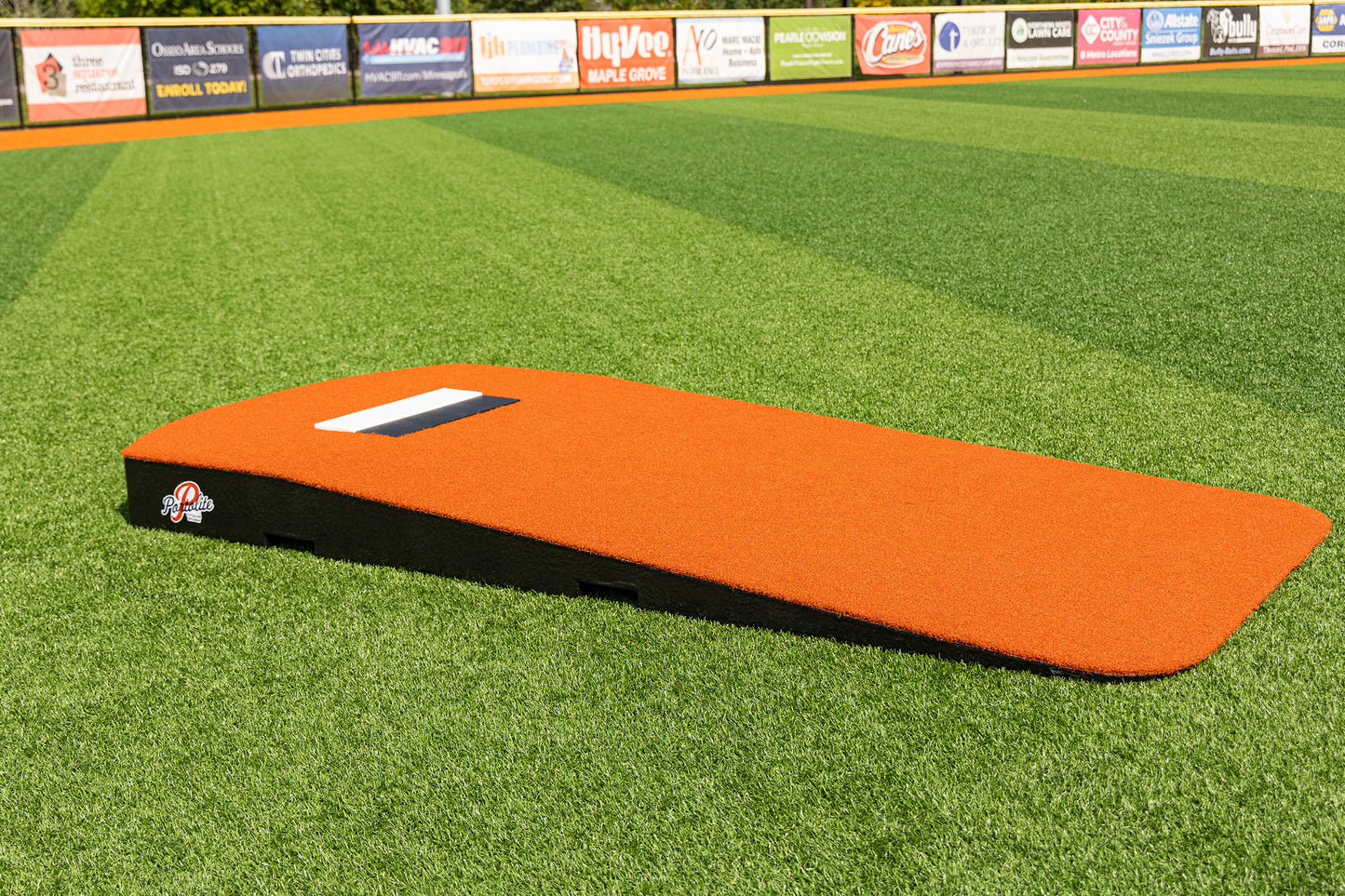 A Portolite Oversized One-Piece Practice Mound in vibrant orange rests on a green artificial turf field, creating an excellent space for pitcher training. Outfield fence advertisements are visible in the background, while the expertly designed mound supports the longest stride length for tall pitchers.
