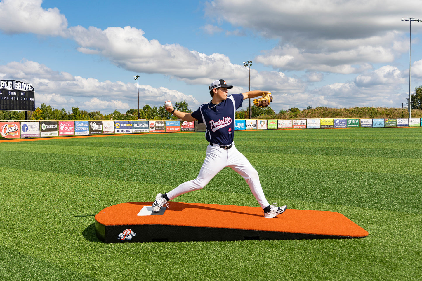 A baseball player in a navy uniform winds up to pitch on the Portolite Oversized One-Piece Practice Mound—designed for the longest stride for tall pitchers—set on a green field, with a scoreboard and advertisements visible in the background under a partly cloudy sky, providing perfect conditions for pitching training.