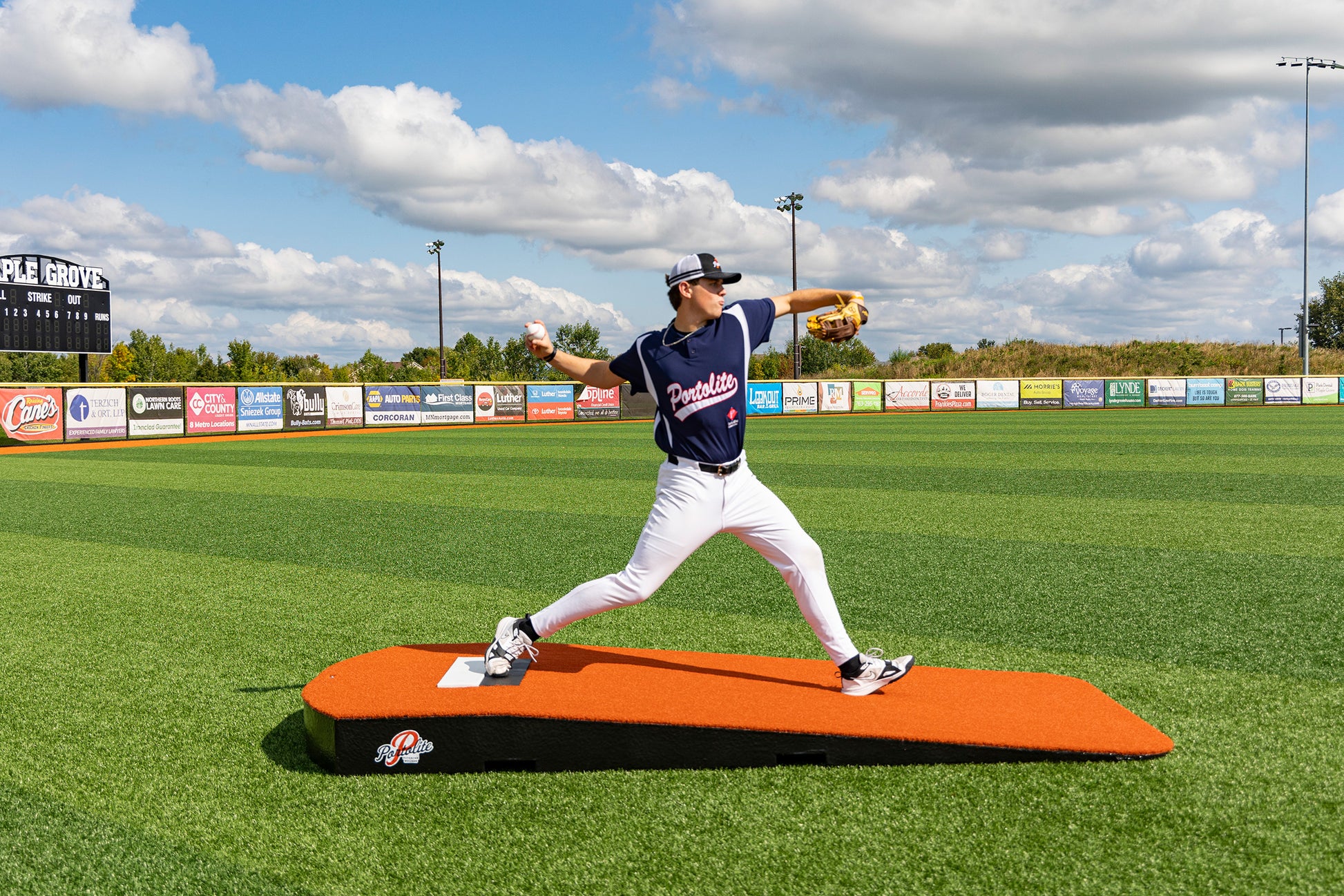 A baseball player in a navy uniform winds up to pitch on the Portolite Oversized One-Piece Practice Mound—designed for the longest stride for tall pitchers—set on a green field, with a scoreboard and advertisements visible in the background under a partly cloudy sky, providing perfect conditions for pitching training.