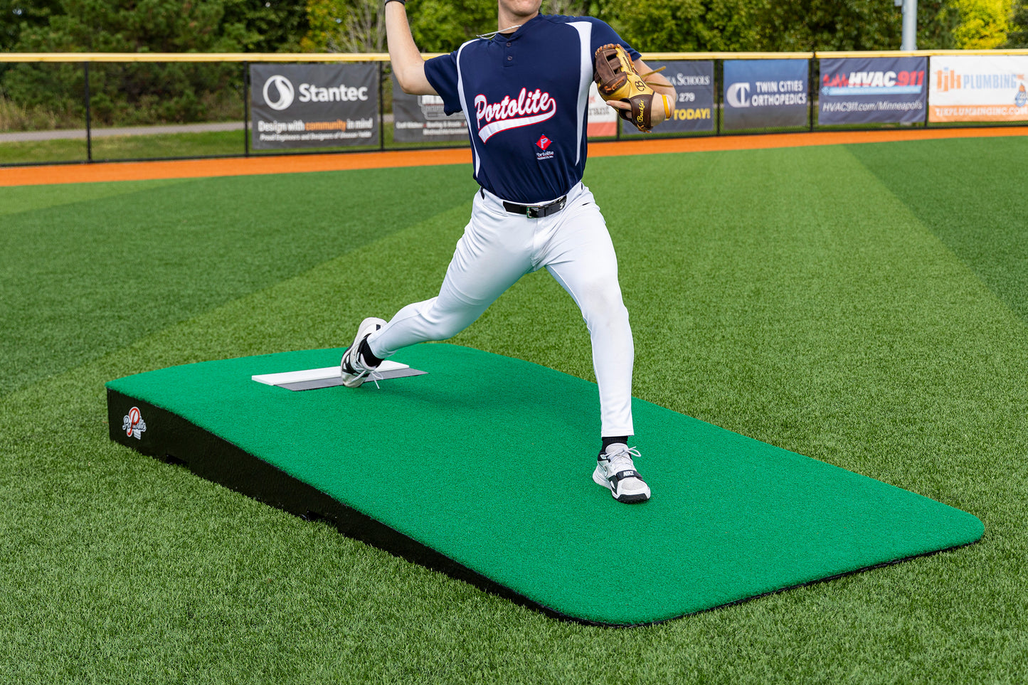 A baseball player in a navy jersey and white pants is pitching from the Portolite Oversized One-Piece Practice Mound | Longest Stride for Tall Pitchers on a turf field, focusing on maximizing stride length during pitcher training, with green grass and advertising banners visible in the background.
