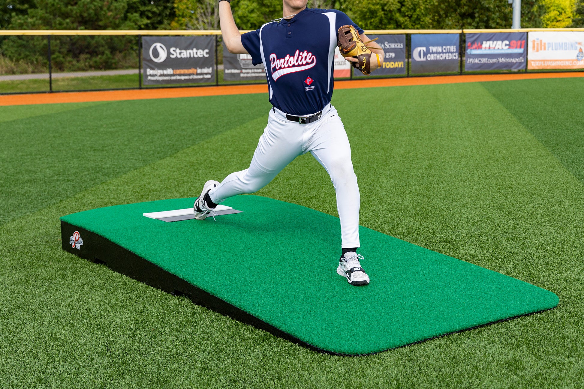 A baseball player in a navy jersey and white pants is pitching from the Portolite Oversized One-Piece Practice Mound | Longest Stride for Tall Pitchers on a turf field, focusing on maximizing stride length during pitcher training, with green grass and advertising banners visible in the background.