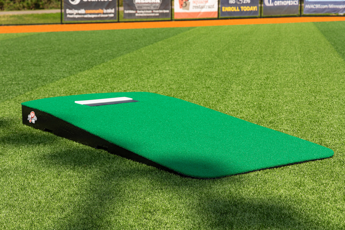 The Portolite Oversized One-Piece Practice Mound, designed to offer the longest stride for tall pitchers, sits on an artificial turf baseball field as a versatile training tool. This green portable pitching mound helps athletes maximize their stride length, while advertisements can be seen on the outfield fence in the background.