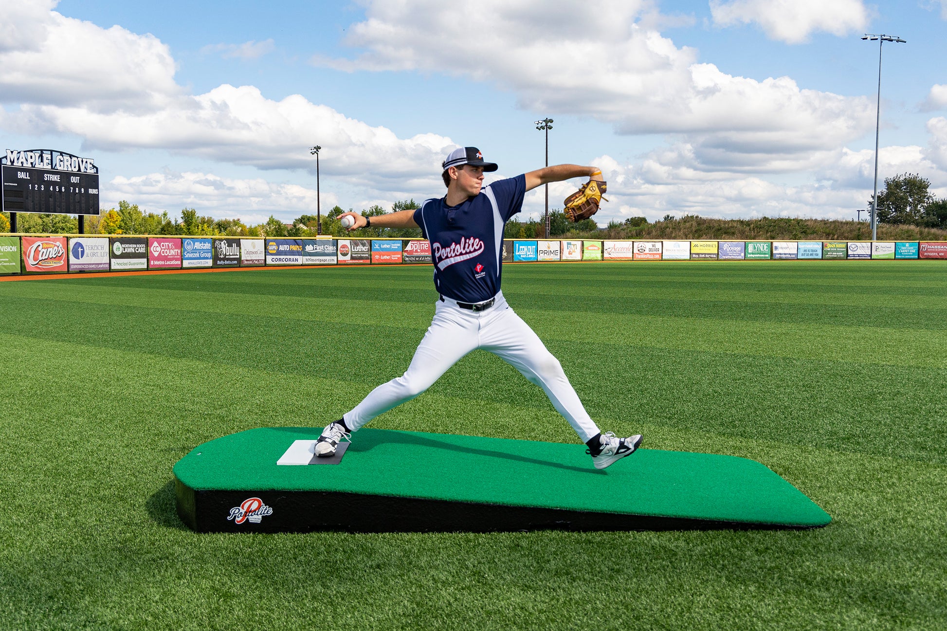 Wearing his uniform, a baseball player throws a pitch from the Portolite Oversized One-Piece Practice Mound | Longest Stride for Tall Pitchers on a field beneath a partly cloudy sky, with the scoreboard and vibrant advertisements in the background emphasizing training for maximum stride length.