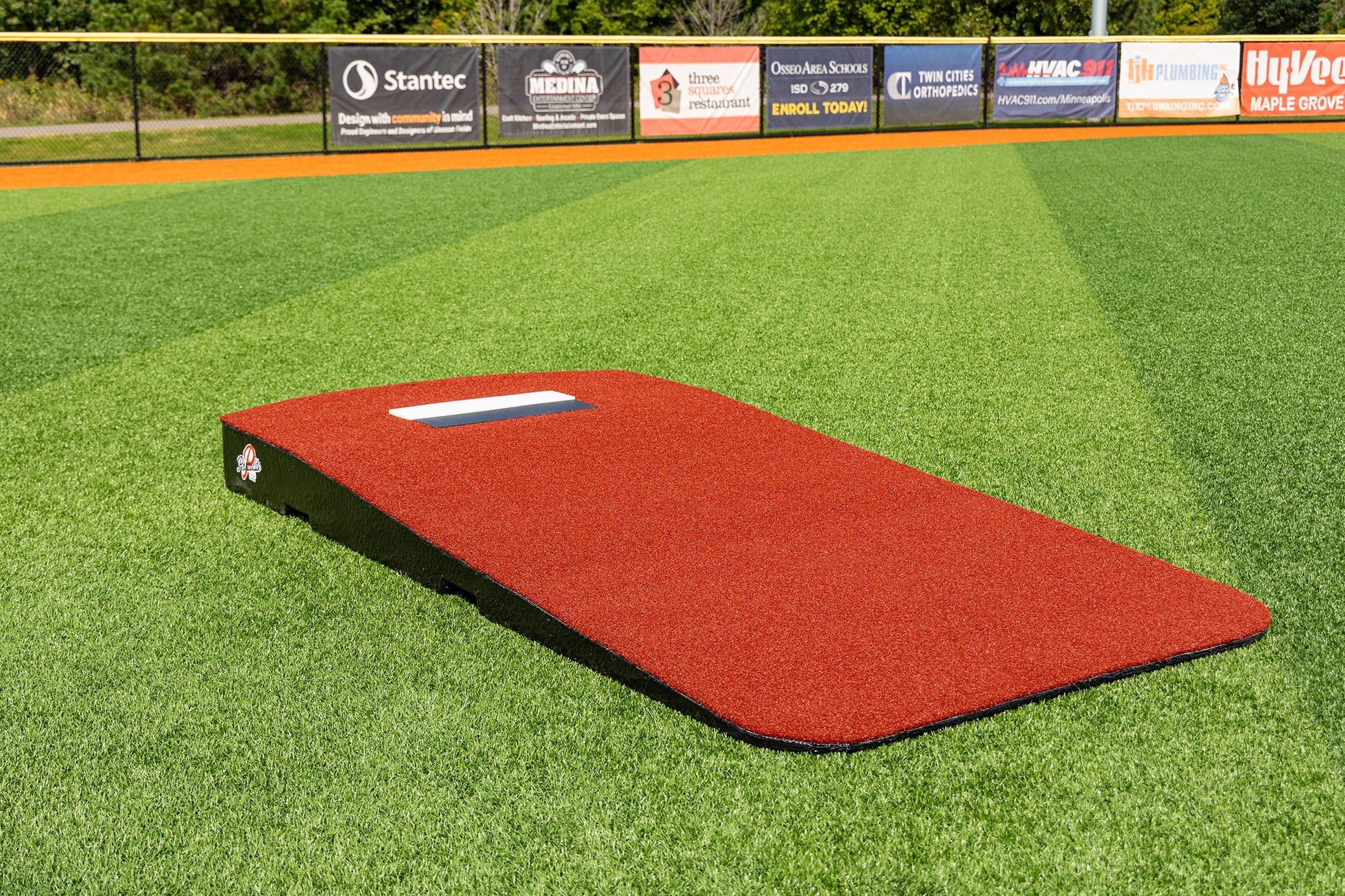 The Portolite Oversized One-Piece Practice Mound | Longest Stride for Tall Pitchers, in a portable red design, sits on artificial turf on a baseball field, offering optimal training space for pitchers to achieve maximum stride length. Outfield fencing and advertisement banners can be seen in the background.