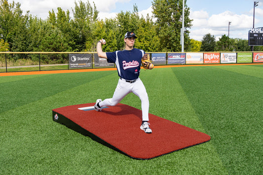 A baseball player in a blue jersey and white pants is pitching from the Portolite Oversized One-Piece Practice Mound | Longest Stride for Tall Pitchers on a green field, demonstrating the longest stride length during pitchers training, with trees, signs, and a scoreboard visible in the background.