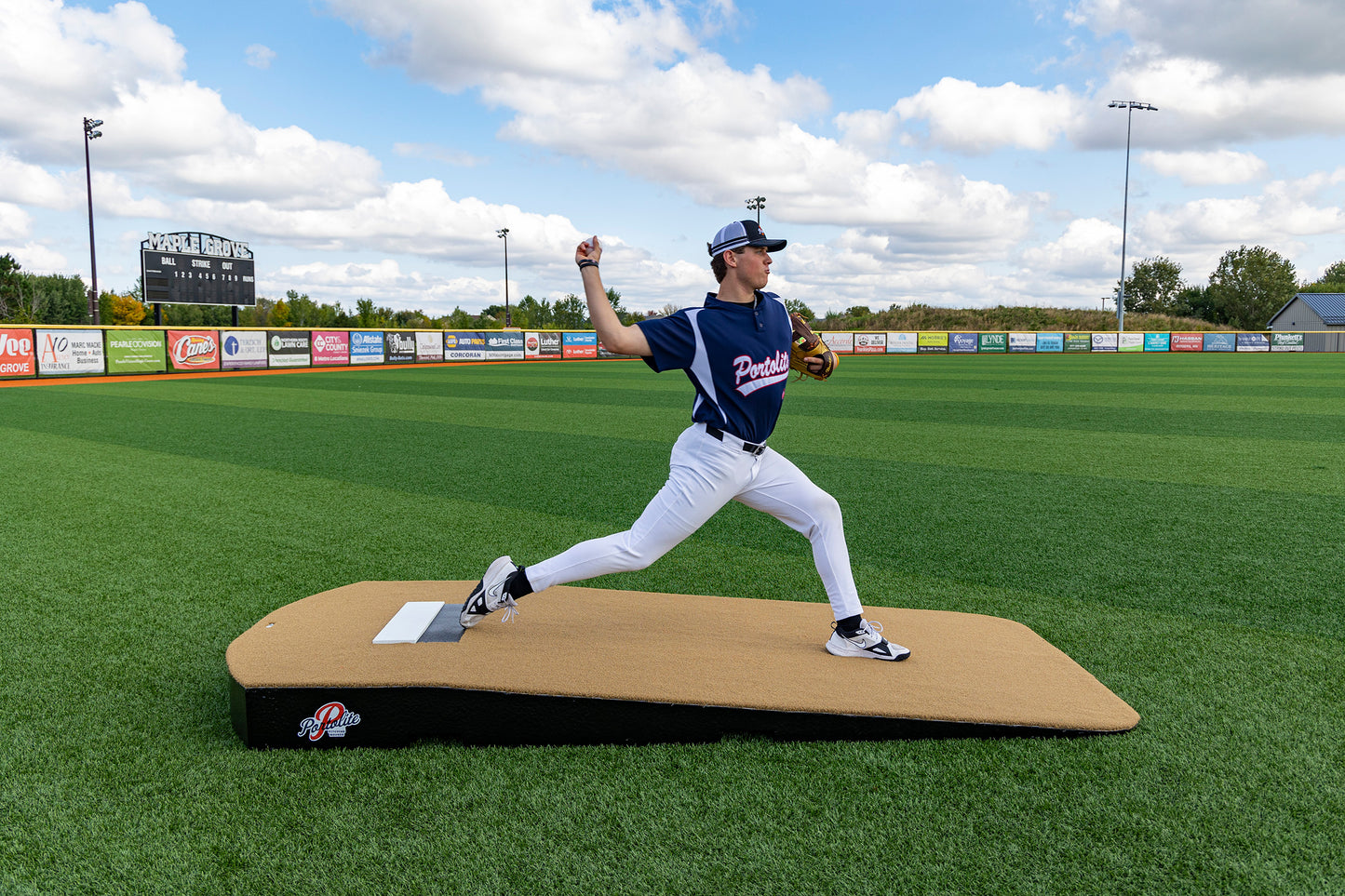 A baseball player in a blue jersey and white pants throws a pitch on the Portolite Oversized One-Piece Practice Mound, designed for the longest stride for tall pitchers, set on a green field with a scoreboard and advertisements visible in the background.