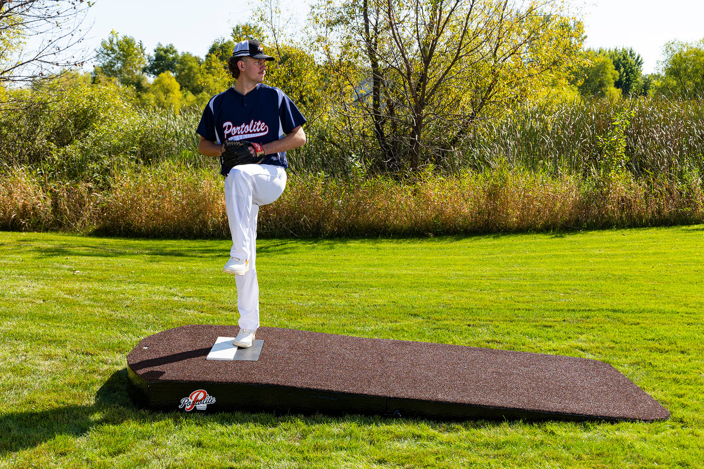A baseball player in uniform stands on the Portolite Oversized Two-Piece Practice Mound, designed with extra width for tall pitchers, preparing to pitch with one leg raised on a grassy outdoor field. Trees and shrubbery are visible in the background under a clear sky.