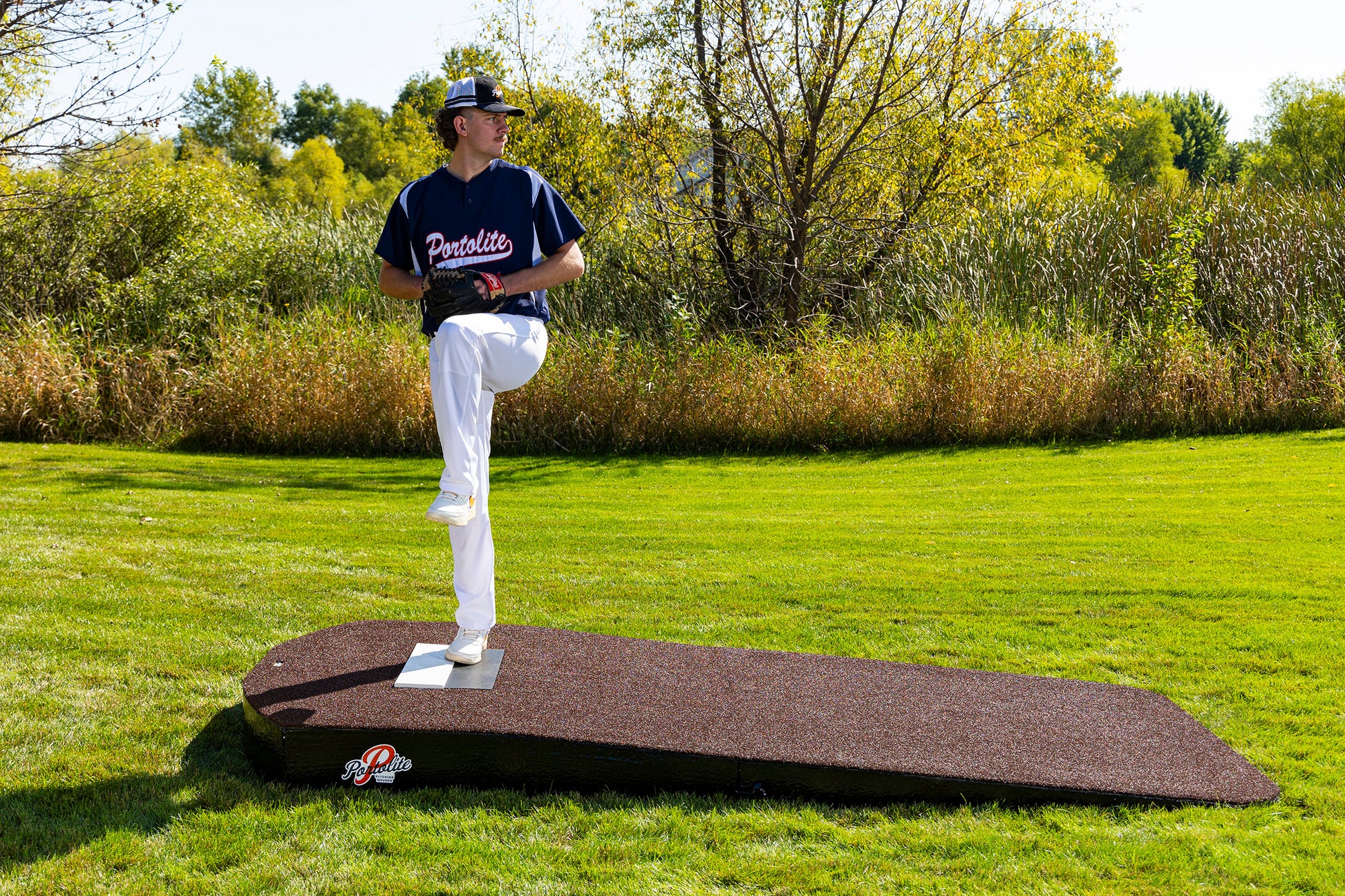 A baseball player in uniform stands on the Portolite Oversized Two-Piece Practice Mound, designed with extra width for tall pitchers, preparing to pitch with one leg raised on a grassy outdoor field. Trees and shrubbery are visible in the background under a clear sky.