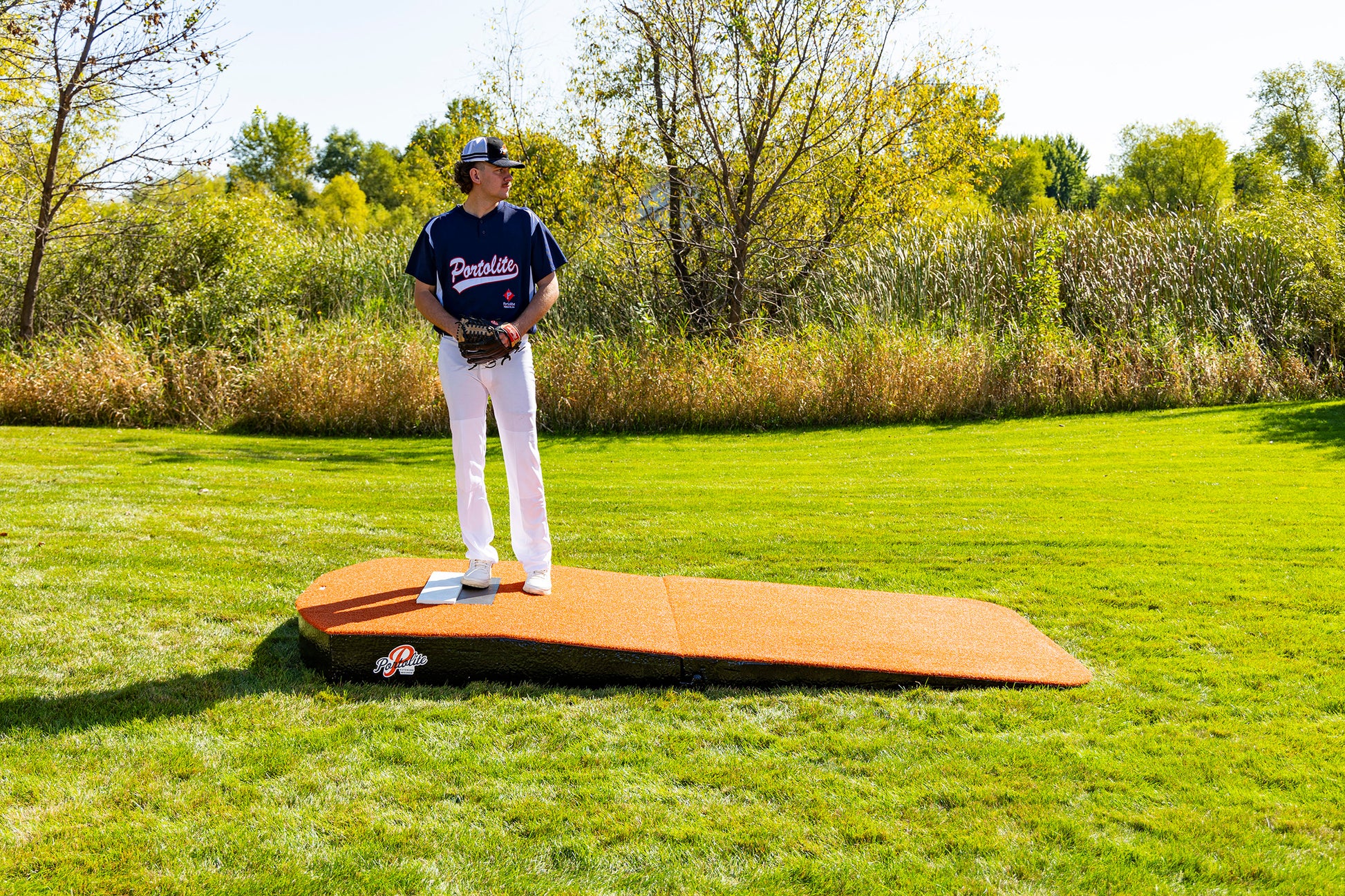 A baseball player wearing a blue jersey and white pants stands on the Portolite Oversized Two-Piece Practice Mound, which features extra width for tall pitchers, positioned on a grassy field with trees and bushes in the background beneath a sunny sky.