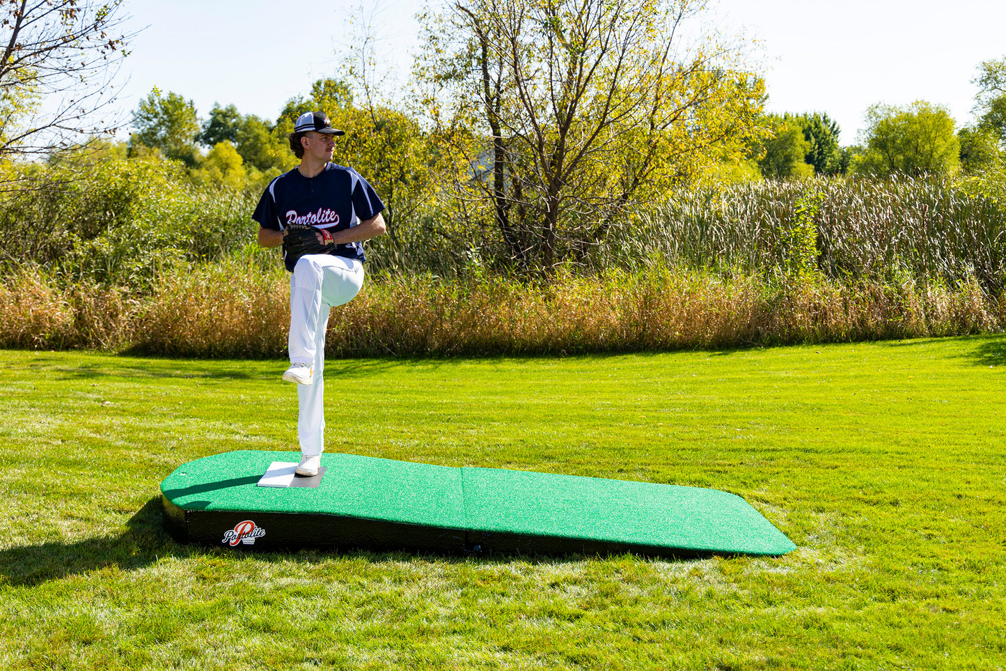 A person wearing a baseball uniform stands on the Portolite Oversized Two-Piece Practice Mound, which offers extra width for tall pitchers, preparing to pitch on a grassy field bordered by trees and bushes.