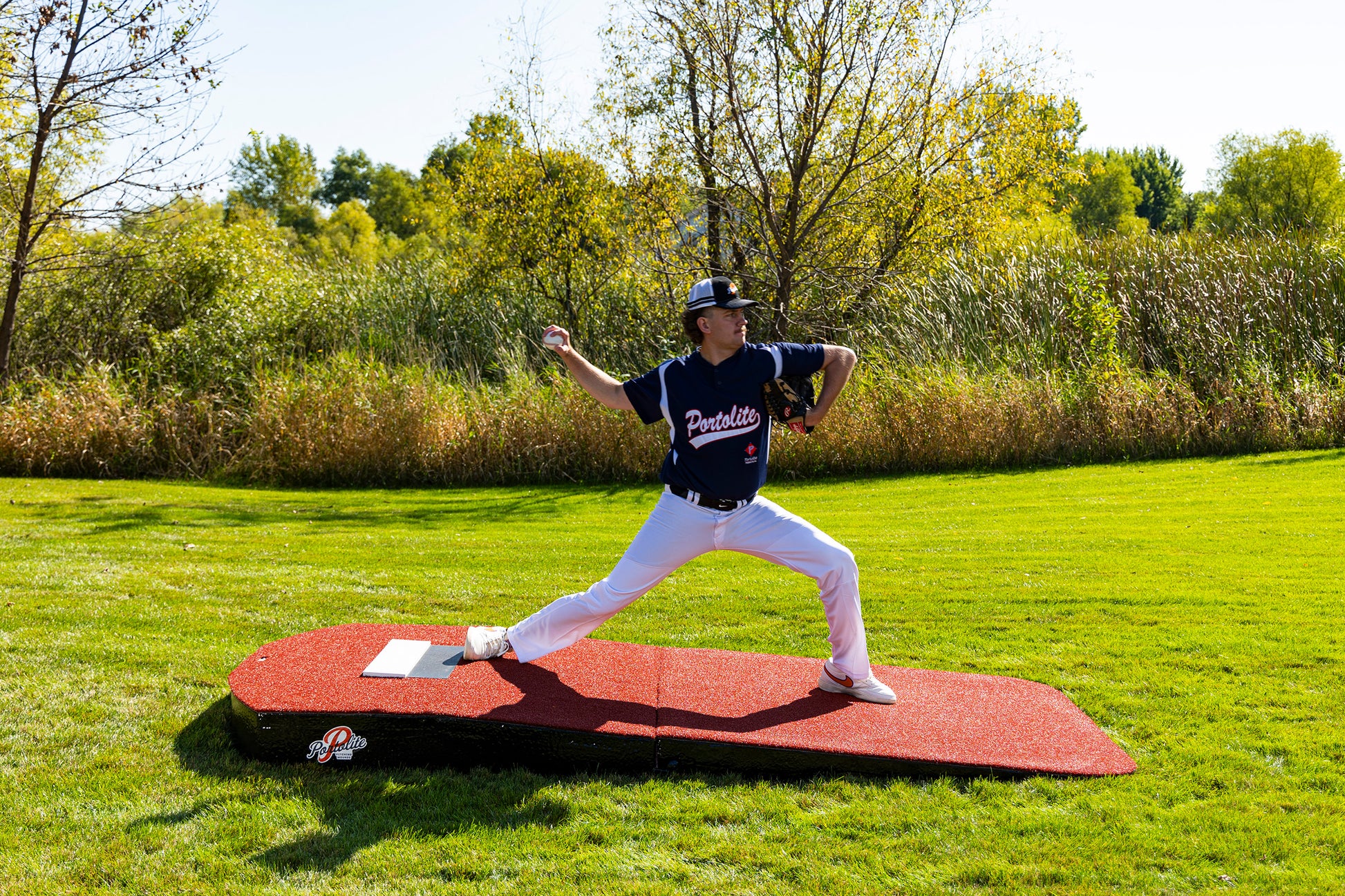 A baseball player in a navy jersey and white pants throws a pitch on a Portolite Oversized Two-Piece Practice Mound, designed with extra width for tall pitchers, set on green grass with trees and bushes in the background under a bright sky.