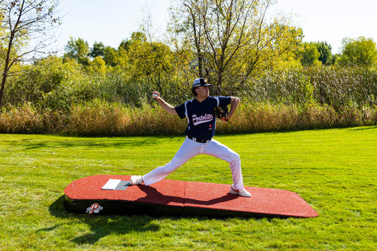 A baseball player in a navy jersey and white pants throws a pitch on a Portolite Oversized Two-Piece Practice Mound, designed with extra width for tall pitchers, set on green grass with trees and bushes in the background under a bright sky.