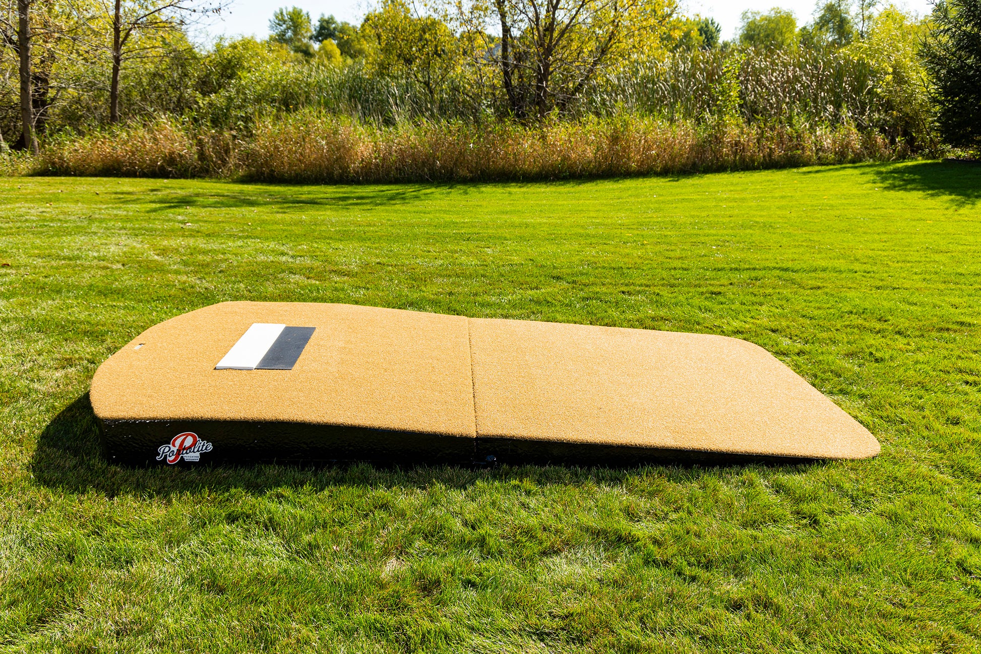 The Portolite Oversized Two-Piece Practice Mound, designed with extra width for tall pitchers, is set on a grassy field surrounded by trees and bushes beneath a sunny sky—ideal for pitching practice or professional mound sessions in any outdoor environment.