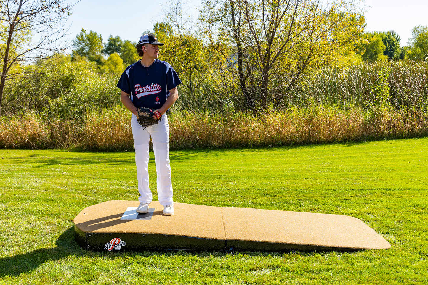 A baseball player in a Portillos jersey stands on a Portolite Oversized Two-Piece Practice Mound, designed with extra width for tall pitchers, in a grassy field with trees and bushes in the background.