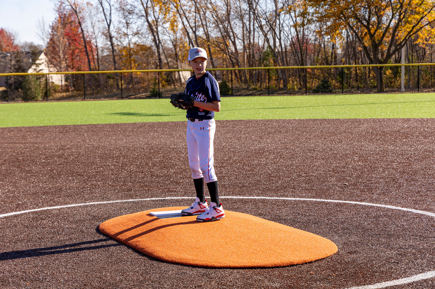 A young Little League baseball player stands poised to pitch on the Portolite 6″ One-Piece Game Mound, designed for ages 8-13, with vibrant autumn trees and sunshine in the background.