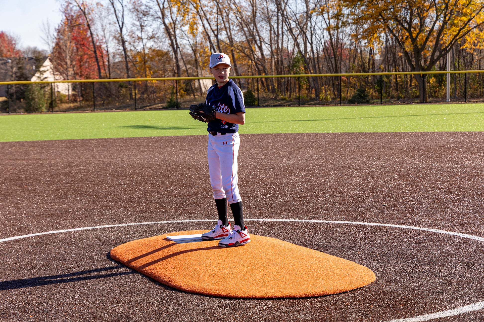 A young Little League baseball player stands poised to pitch on the Portolite 6″ One-Piece Game Mound, designed for ages 8-13, with vibrant autumn trees and sunshine in the background.