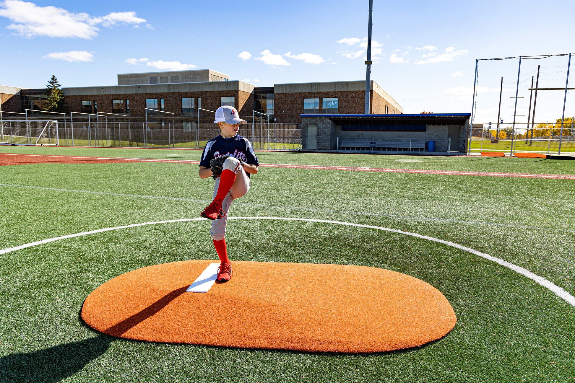 A young Little League player, age 8-13, stands mid-windup on the Portolite 6″ One-Piece Game Mound at an outdoor baseball field, with a school building and dugout visible in the background.