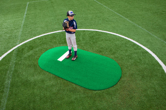 A young Little League baseball player stands on the Portolite 6″ One-Piece Game Mound, designed for ages 8-13, wearing a blue uniform and cap, holding a glove and looking to the side on a green field.