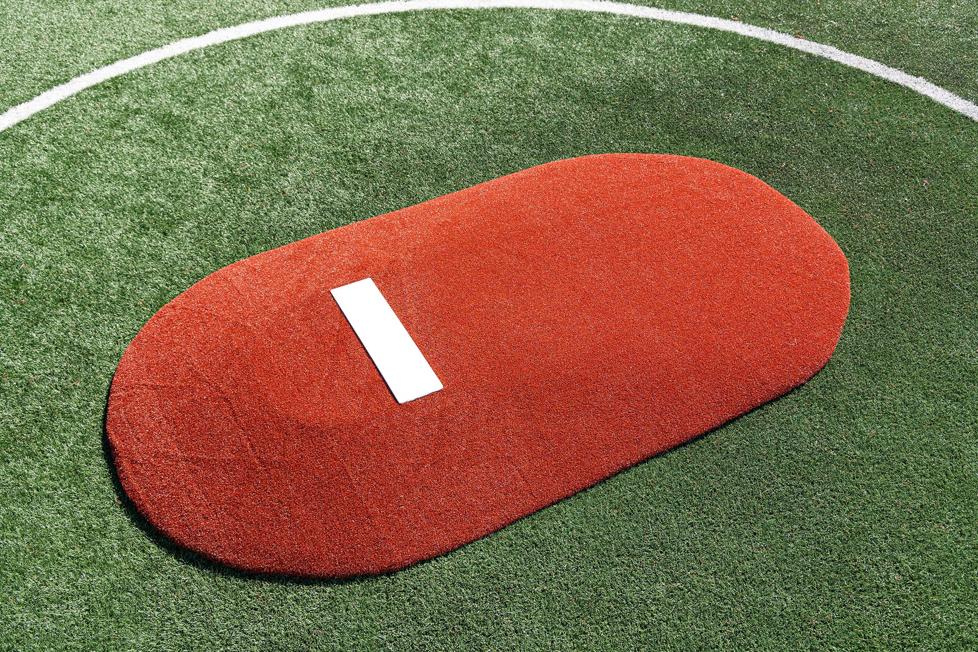 The Portolite 6″ One-Piece Game Mound for ages 8-13, featuring an artificial red surface and a white rubber plate, rests on green turf outlined by a white curved line—ideal for Little League tournament play.