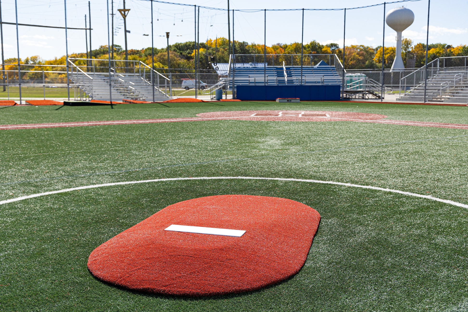 A baseball field featuring the Portolite 6″ One-Piece Game Mound for ages 8-13 in the foreground, with home plate and stands in the background, and trees showing autumn colors beyond the fence—ideal for Little League tournament play.
