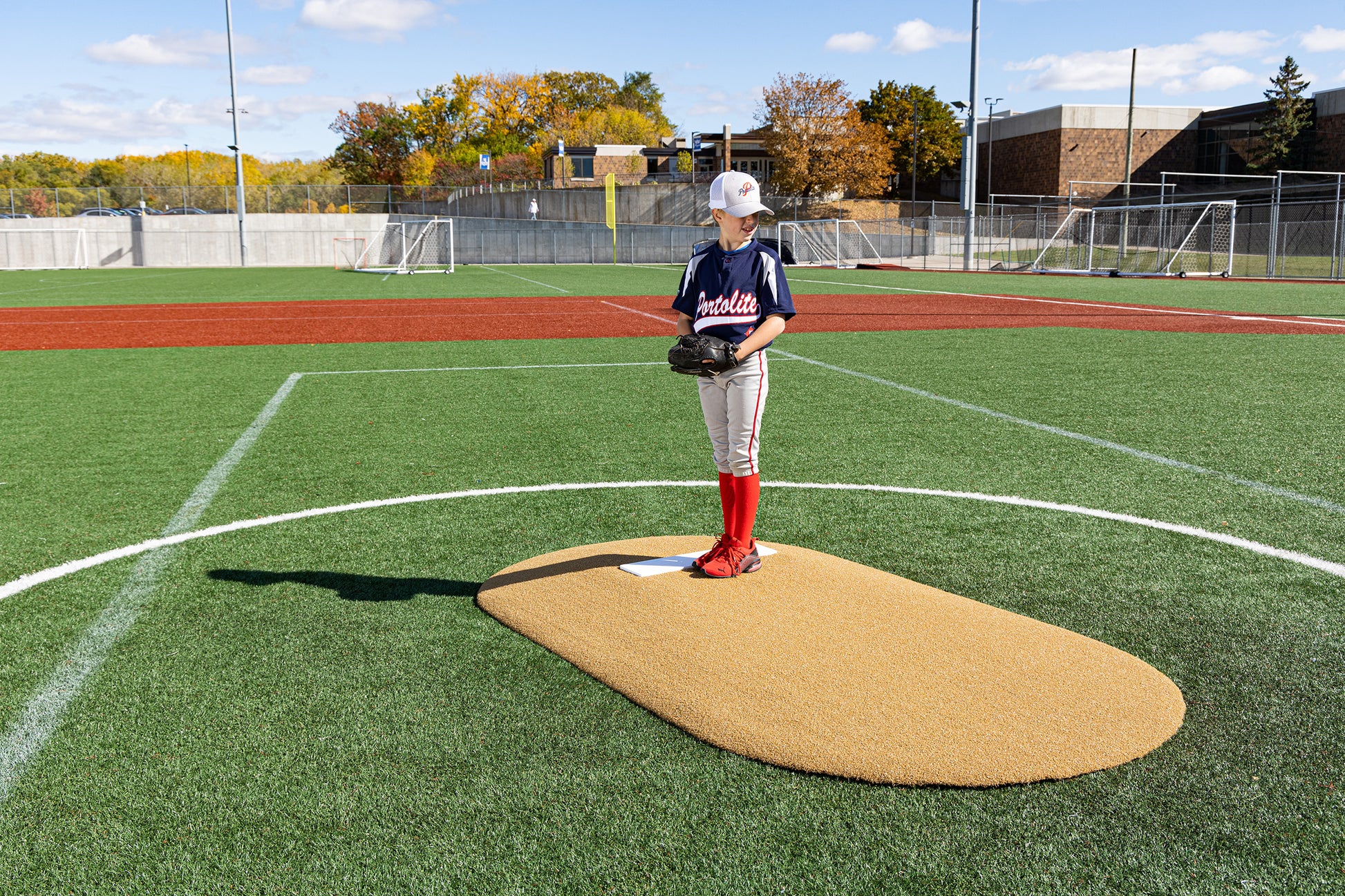 A youth baseball player stands on the Portolite 6″ One-Piece Game Mound, ready with a glove and dressed in a blue jersey, red socks, and a cap on a sunny day at a Little League field for ages 8-13.