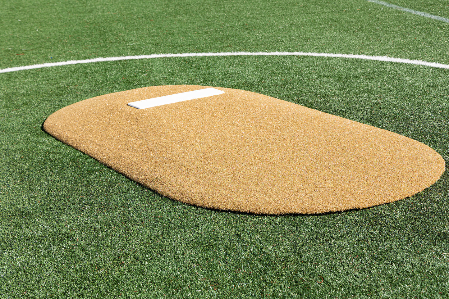 The Portolite 6″ One-Piece Game Mound for ages 8-13 features a tan artificial turf surface atop a green synthetic field, complete with a white rectangular pitcher’s rubber and a curved white line in the background—ideal for Little League games or tournament play.
