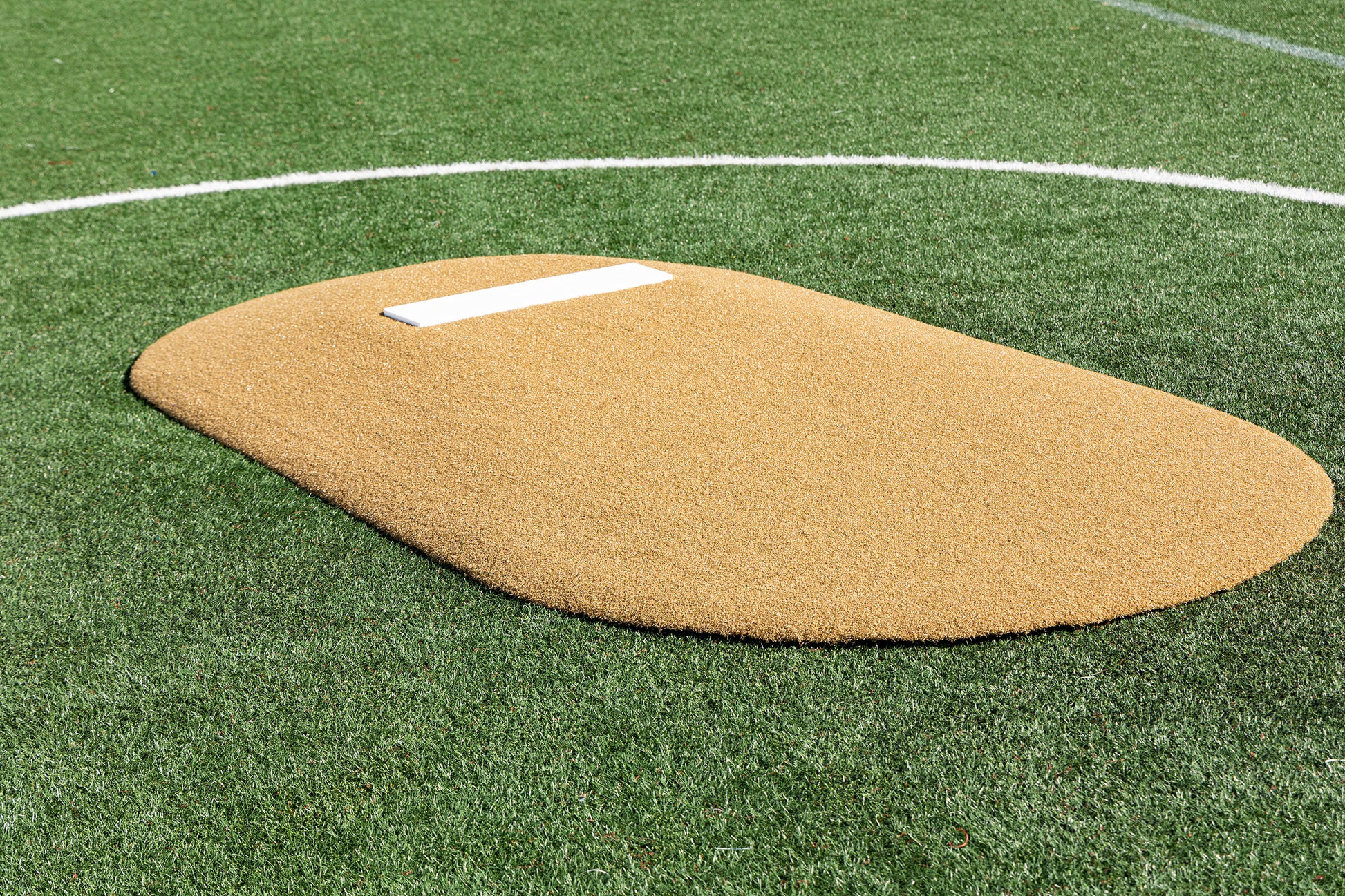 The Portolite 6″ One-Piece Game Mound for ages 8-13 features a tan artificial turf surface atop a green synthetic field, complete with a white rectangular pitcher’s rubber and a curved white line in the background—ideal for Little League games or tournament play.