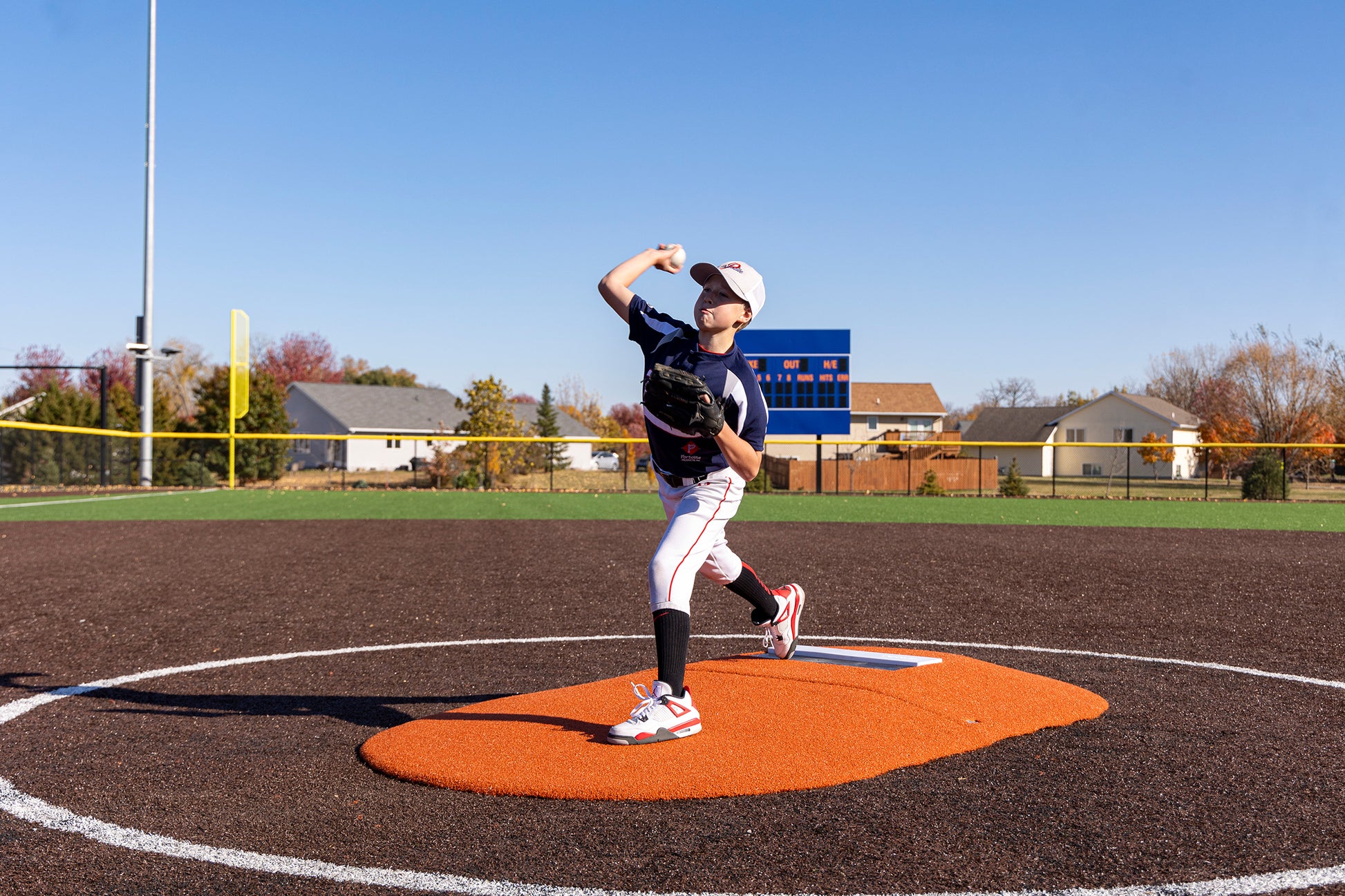 A young baseball player in uniform is pitching on the Portolite 6″ Two-Piece Game Mound for ages 9-13 at a baseball field under a clear blue sky, with houses and trees in the background.