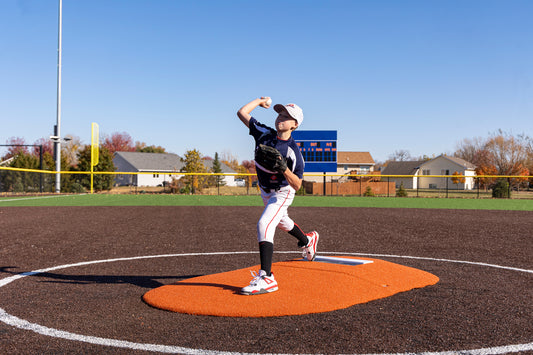 A young baseball player in uniform is pitching on the Portolite 6″ Two-Piece Game Mound for ages 9-13 at a baseball field under a clear blue sky, with houses and trees in the background.