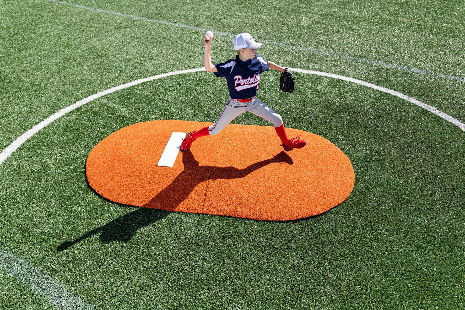 A young baseball pitcher in a blue Panthers jersey and red socks throws a ball from a Portolite 6″ Two-Piece Game Mound, designed for ages 9-13, set on a green field, casting a long shadow.