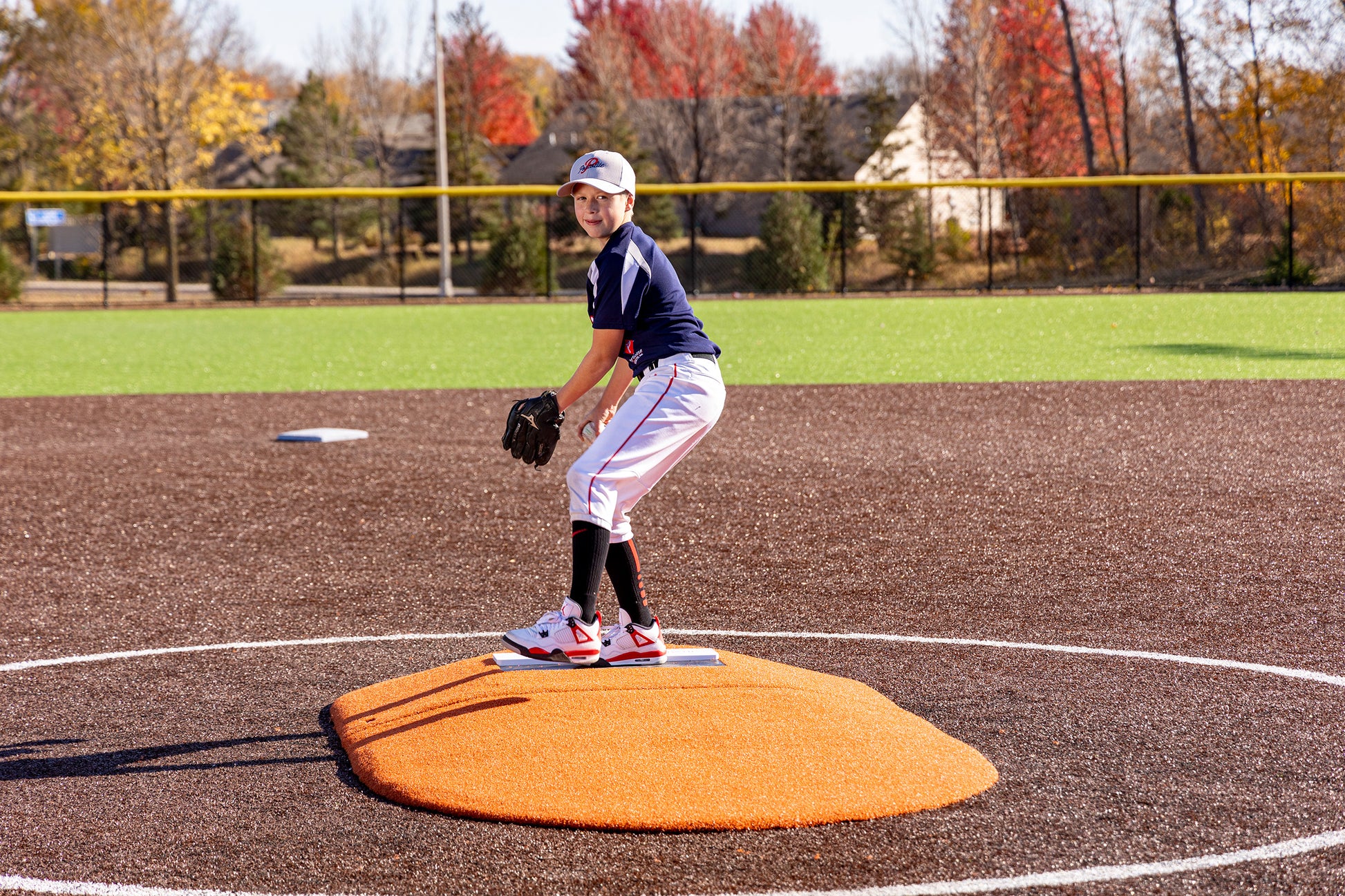 A young baseball player stands on the Portolite 6″ Two-Piece Game Mound, designed for ages 9-13, poised to pitch on a field with brown turf and green grass. Trees with autumn leaves are visible in the background.