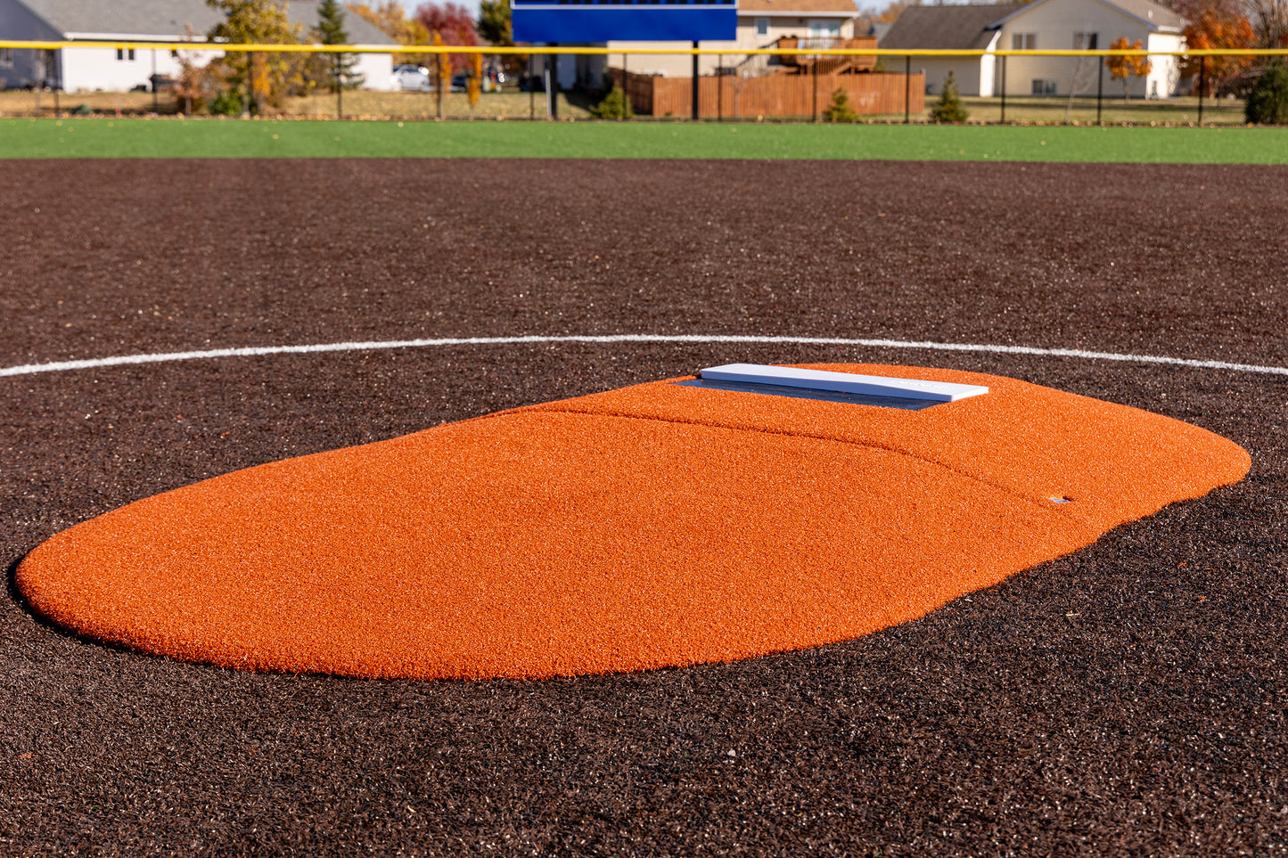 Introducing the Portolite 6″ Two-Piece Game Mound, designed for ages 9-13—an artificial turf portable pitching mound featuring a bright orange surface and a white rubber plate. Set it on a brown infield with lush green grass and neighborhood houses in the background, making it perfect for tournament play or as your go-to game mound.