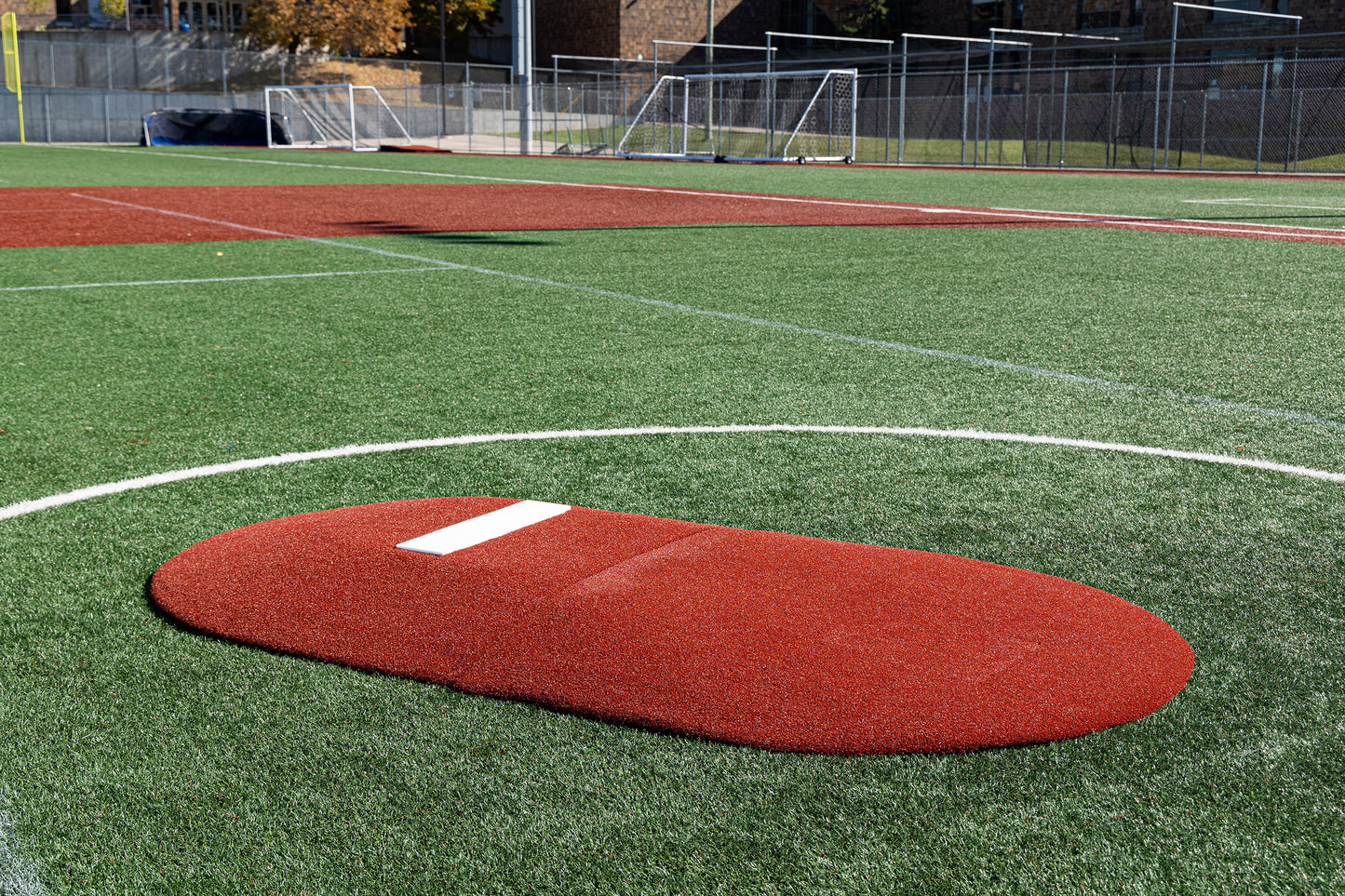 The Portolite 6″ Two-Piece Game Mound, designed for ages 9-13, in vibrant red with a white rectangular rubber, is positioned on a green turf baseball field—perfect for tournament play. In the background, you can see soccer goals and a chain-link fence.