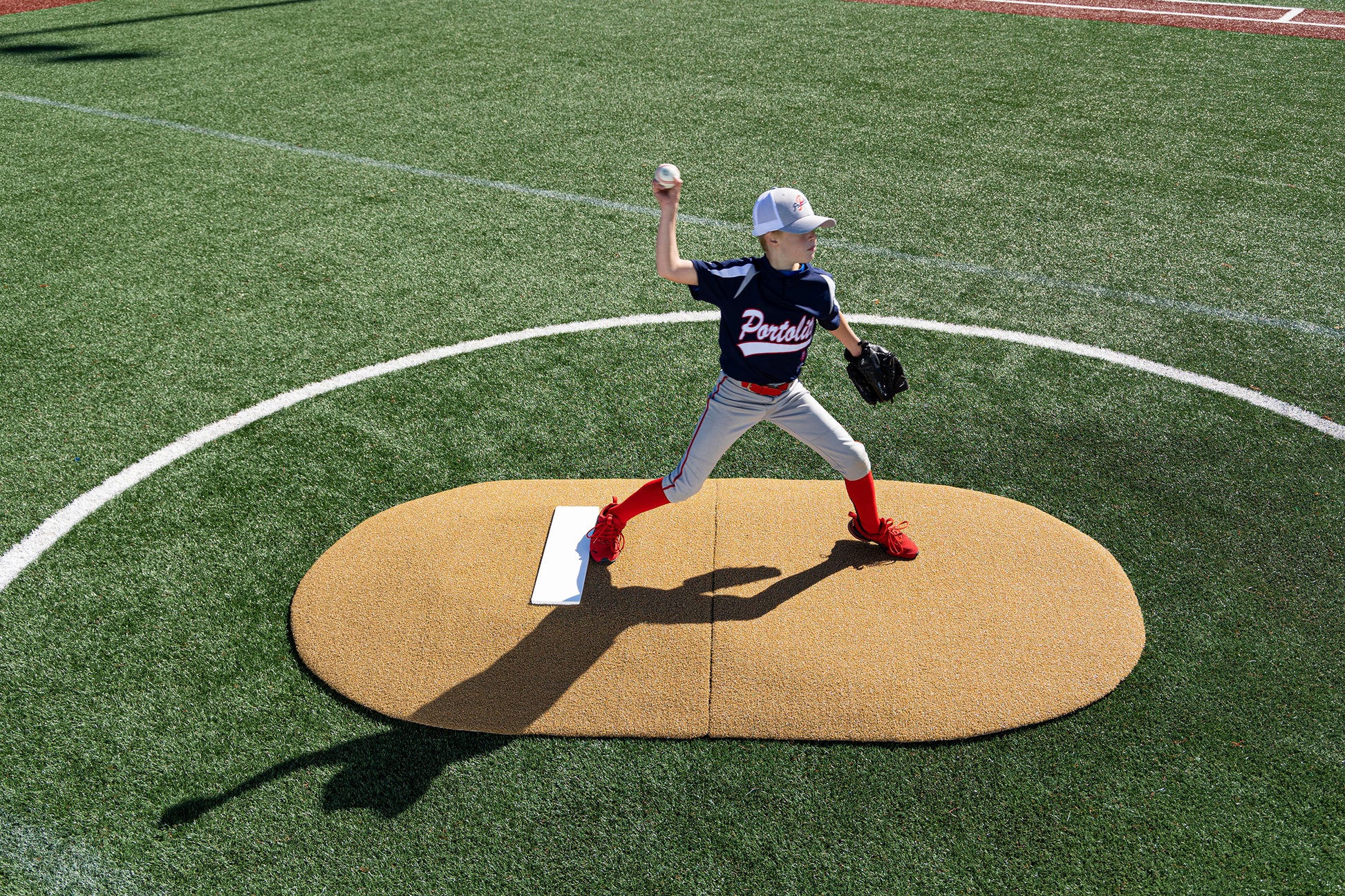 A young baseball player in a navy jersey and red socks is pitching mid-throw on the Portolite 6″ Two-Piece Game Mound (for ages 9–13), set atop a green artificial turf field.