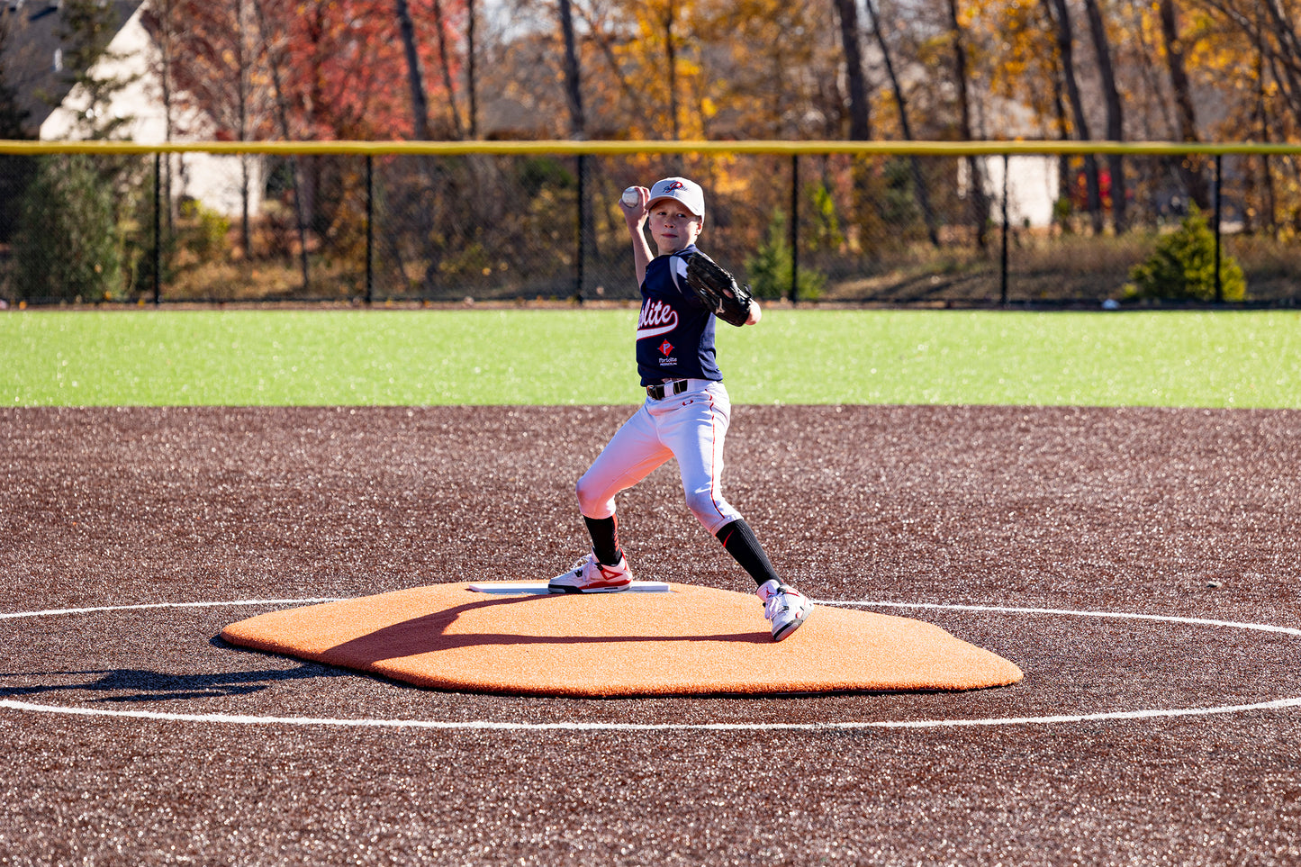 A young little league baseball player in uniform winds up to pitch on the Portolite 6″ Oversized One-Piece Game Mound, designed for ages 8-13, on an outdoor field with autumn trees and a yellow fence in the background.