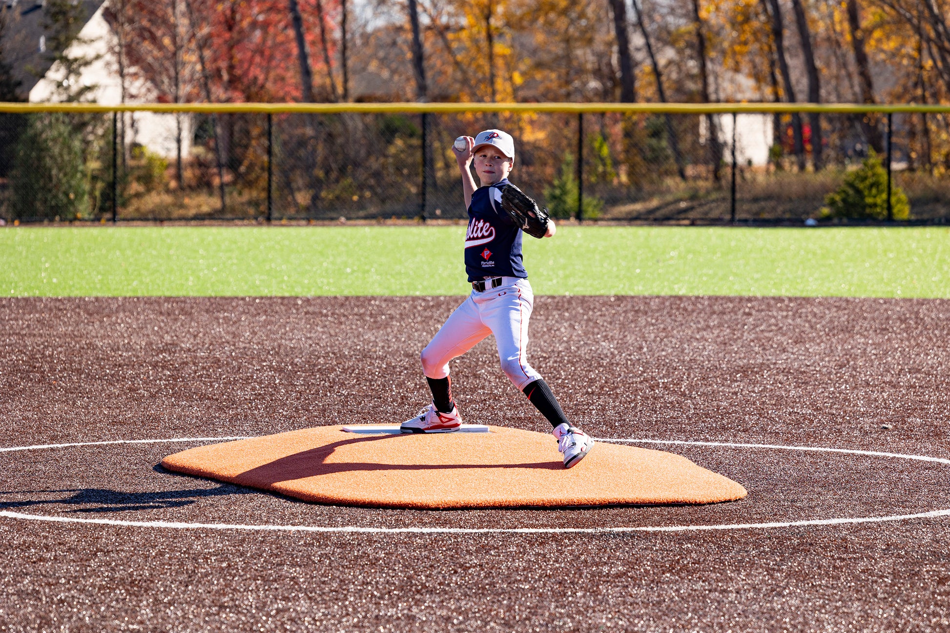 A young little league baseball player in uniform winds up to pitch on the Portolite 6″ Oversized One-Piece Game Mound, designed for ages 8-13, on an outdoor field with autumn trees and a yellow fence in the background.