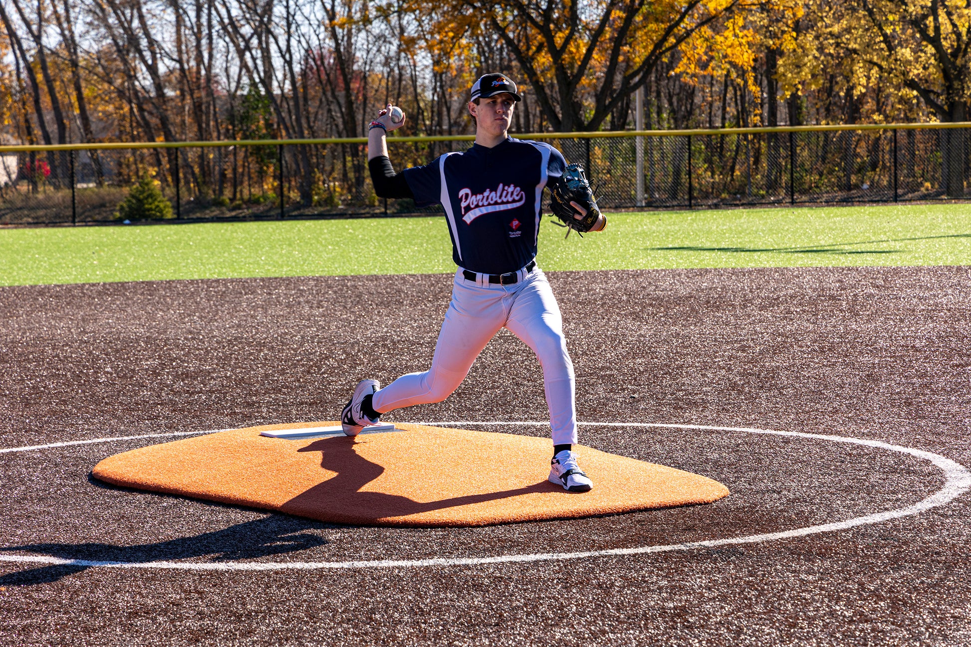 A baseball player in a blue Penfield jersey is demonstrating impressive pitching mechanics on a Portolite 6″ Oversized One-Piece Game Mound (for ages 8-13) during a sunny day, with autumn trees in the background.