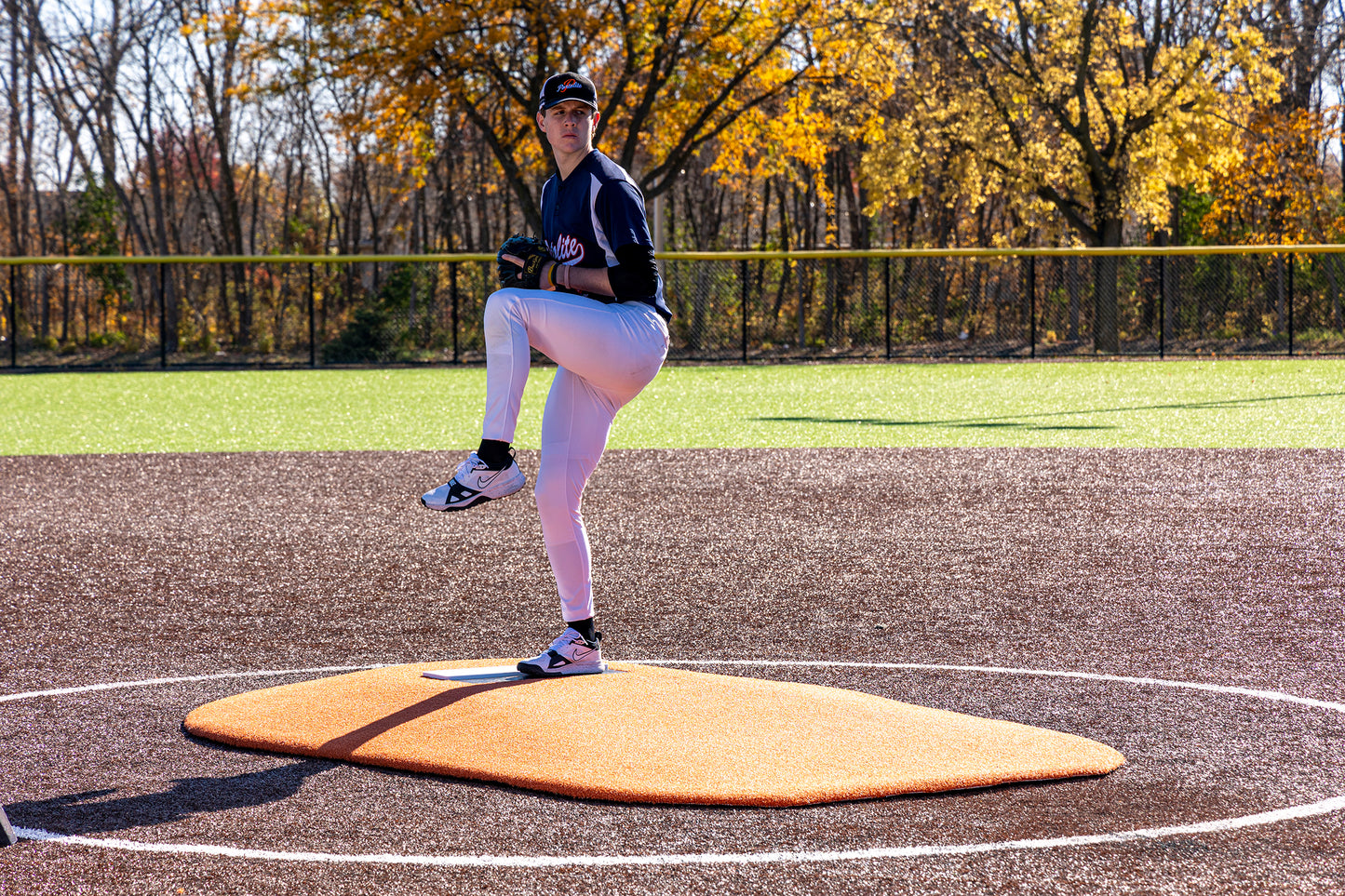 A young baseball player, dressed in a dark jersey and white pants, demonstrates precise pitching mechanics from atop the Portolite 6″ Oversized One-Piece Game Mound (for ages 8-13), with vibrant autumn foliage visible in the background.