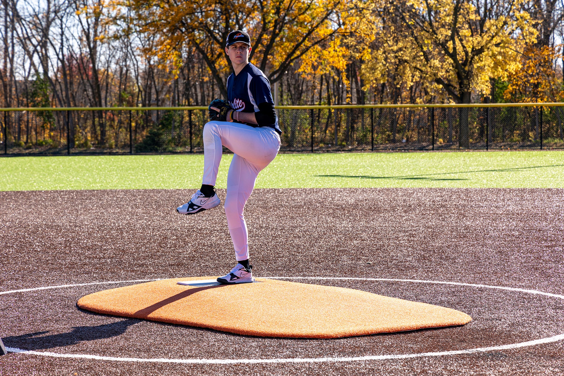 A young baseball player, dressed in a dark jersey and white pants, demonstrates precise pitching mechanics from atop the Portolite 6″ Oversized One-Piece Game Mound (for ages 8-13), with vibrant autumn foliage visible in the background.
