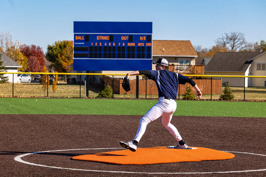 A baseball pitcher in a navy jersey and white pants winds up to throw a pitch from the Portolite 6″ Oversized Two-Piece Game Mound for ages 10-14, set on an outdoor field with a blue scoreboard and houses in the background, highlighting the enhanced setup often seen during tournament play.