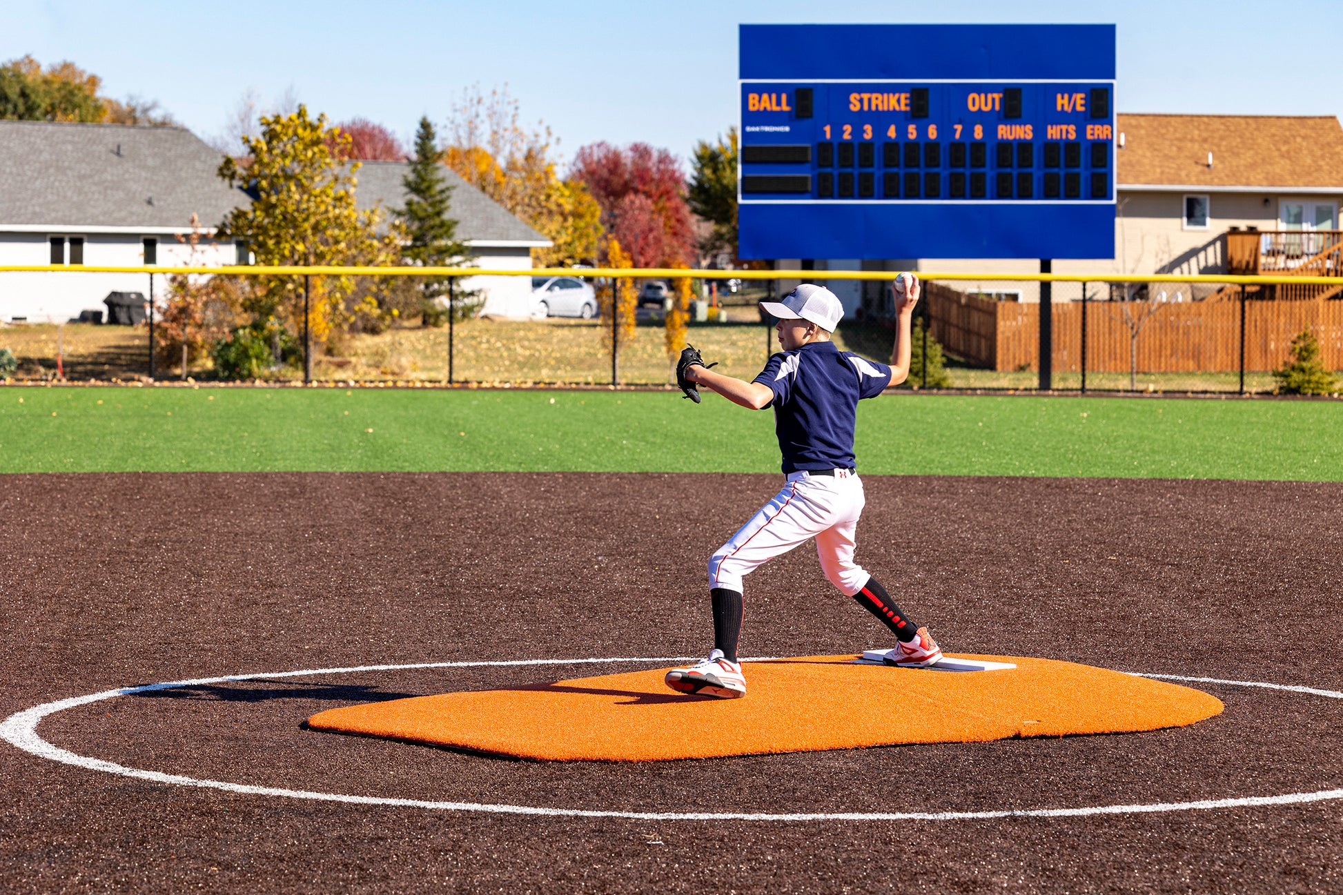 A young pitcher in a navy uniform winds up to throw from the Portolite 6″ Oversized Two-Piece Game Mound, designed for ages 10-14. A blue scoreboard stands in the background on a sunny fall day, highlighting the intensity of tournament play.