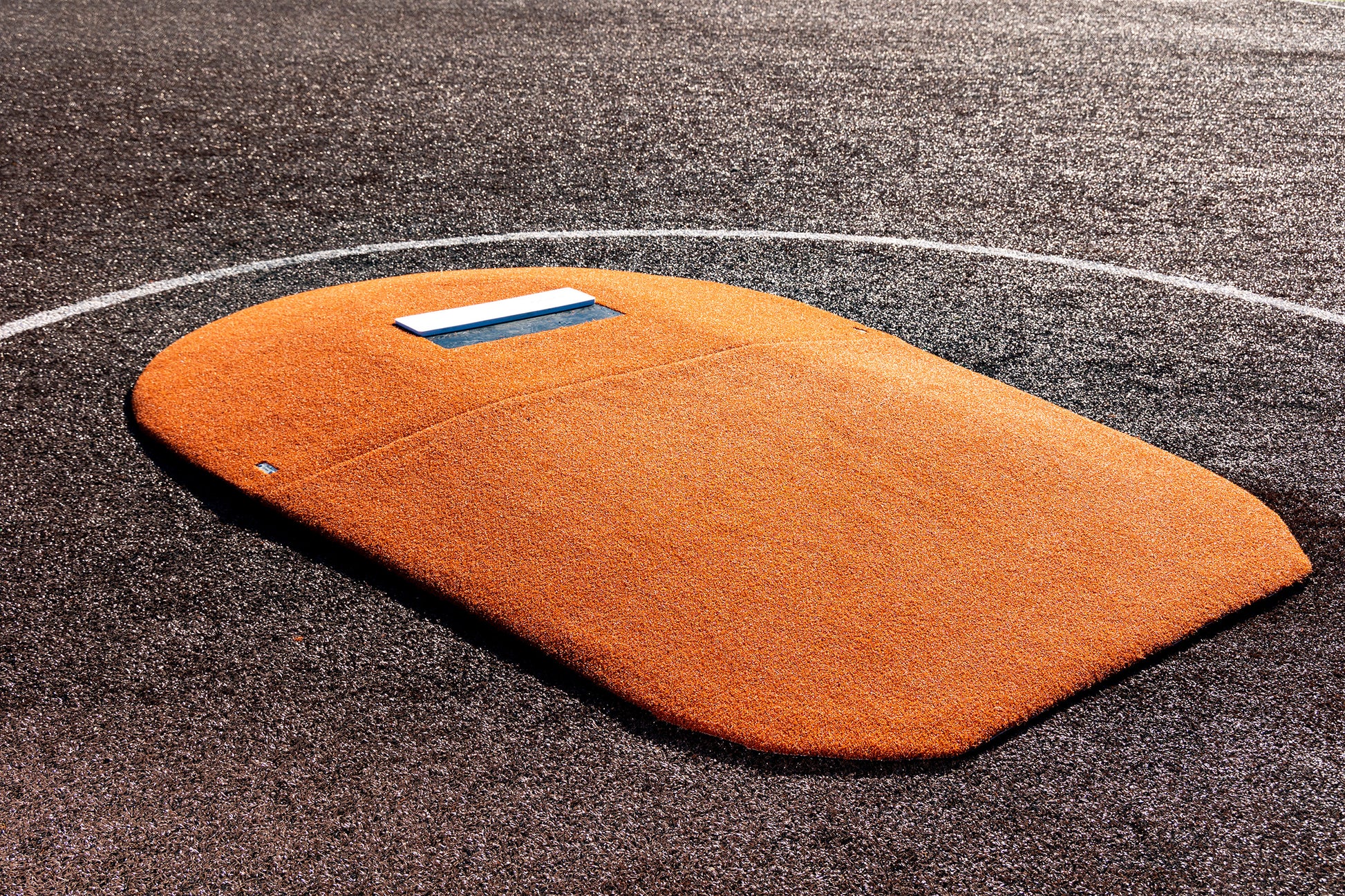 The Portolite 6″ Oversized Two-Piece Game Mound for ages 10-14, featuring an orange portable design with a white rubber pitching plate, is set on a dark synthetic turf baseball field with a white chalk circle—making it ideal for tournament play.