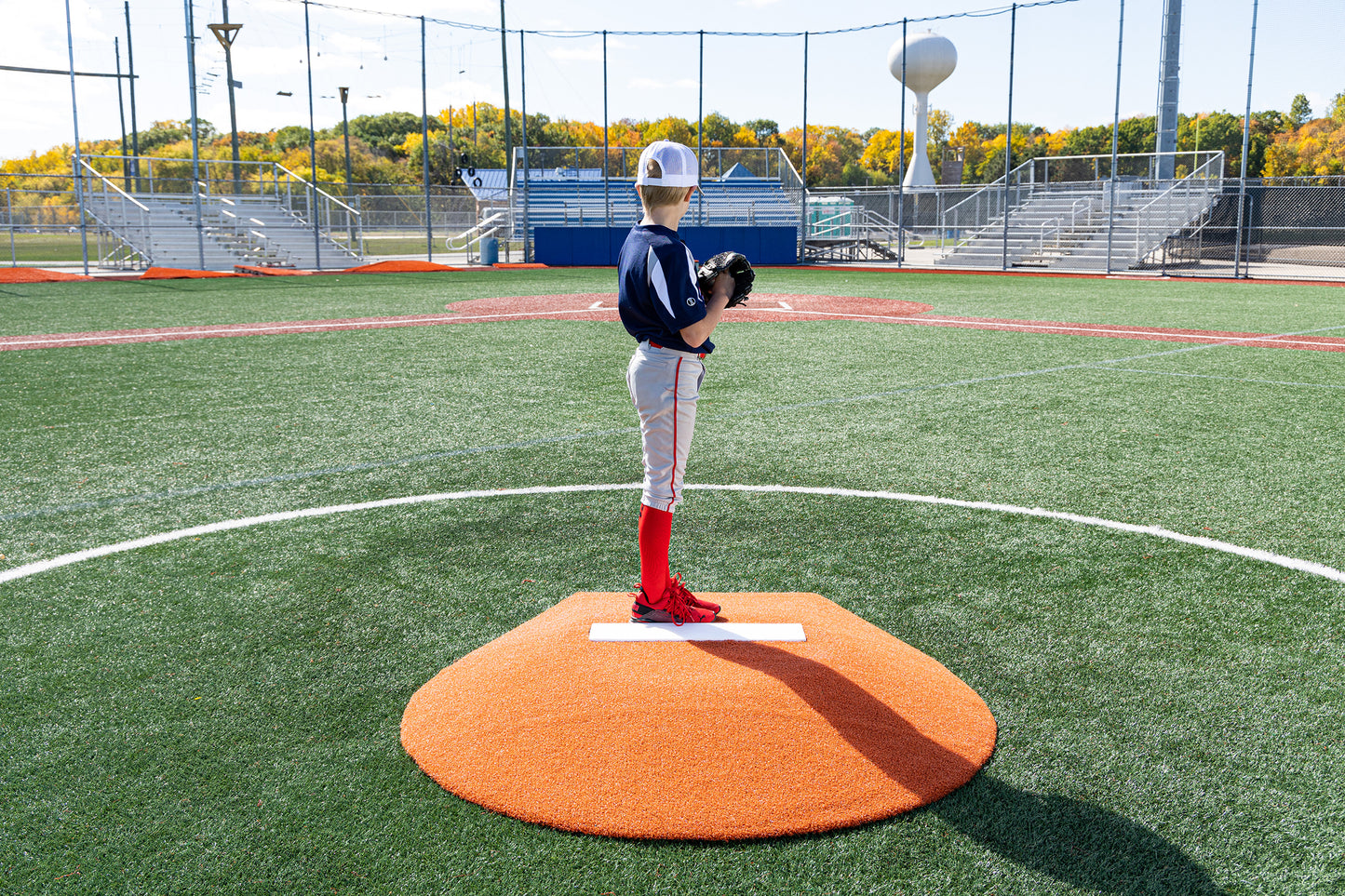 A young Little League baseball player in a navy jersey, white pants, and red socks stands confidently on the Portolite 6″ Stride Off Game Mound (for ages 13 & under), holding a glove while facing an empty field with bleachers and fall trees in the background.